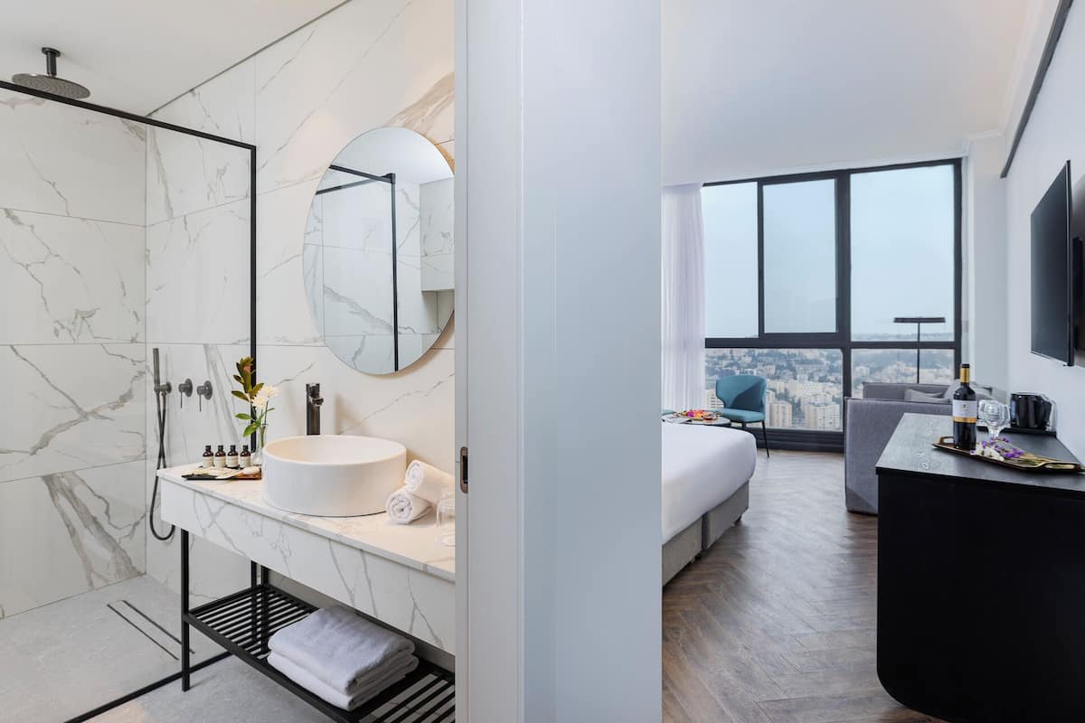 A modern bathroom with a marble vanity, a large mirror, and a shower. The room features a large window with a city view.