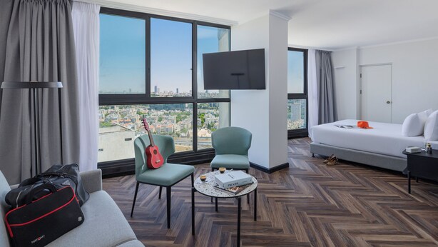 A hotel room with a large window, a flat-screen TV, a bed, a sofa, two armchairs, a coffee table, and a guitar. - Brown Jerusalem Hotel & Convention Center (Jerusalem)
