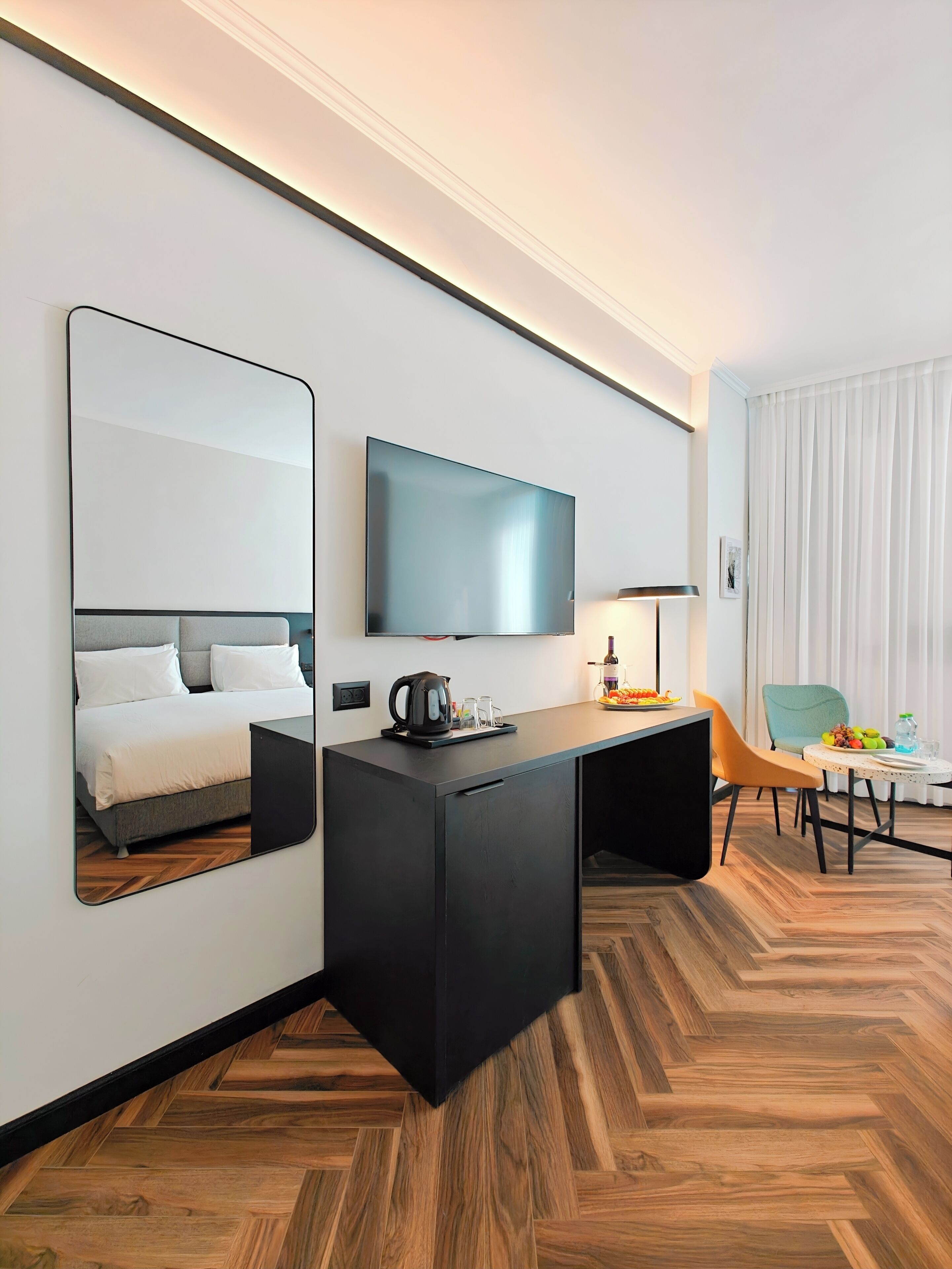 A modern hotel room with a large bed, a flat-screen TV mounted on the wall, a desk with a kettle, and a dining area with a table and chairs.