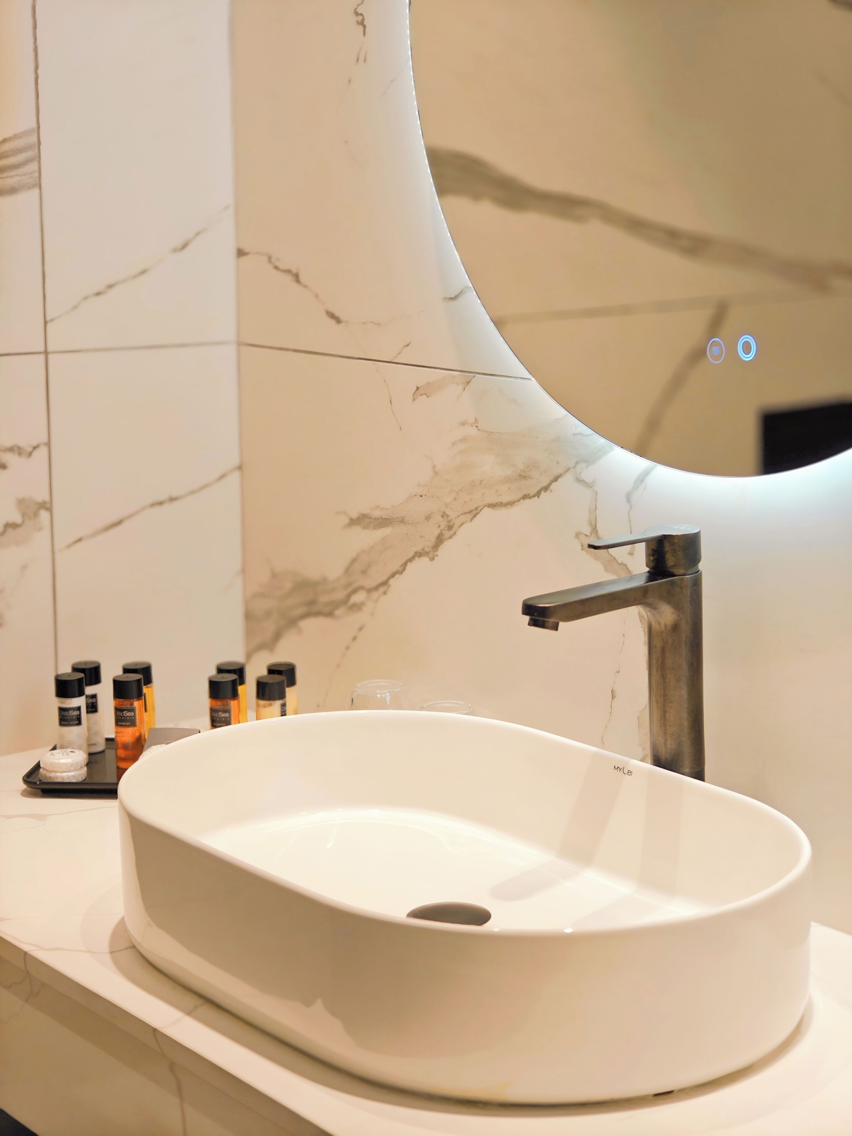 A bathroom with a white sink, a modern faucet, and a curved mirror. There are toiletries on the counter.