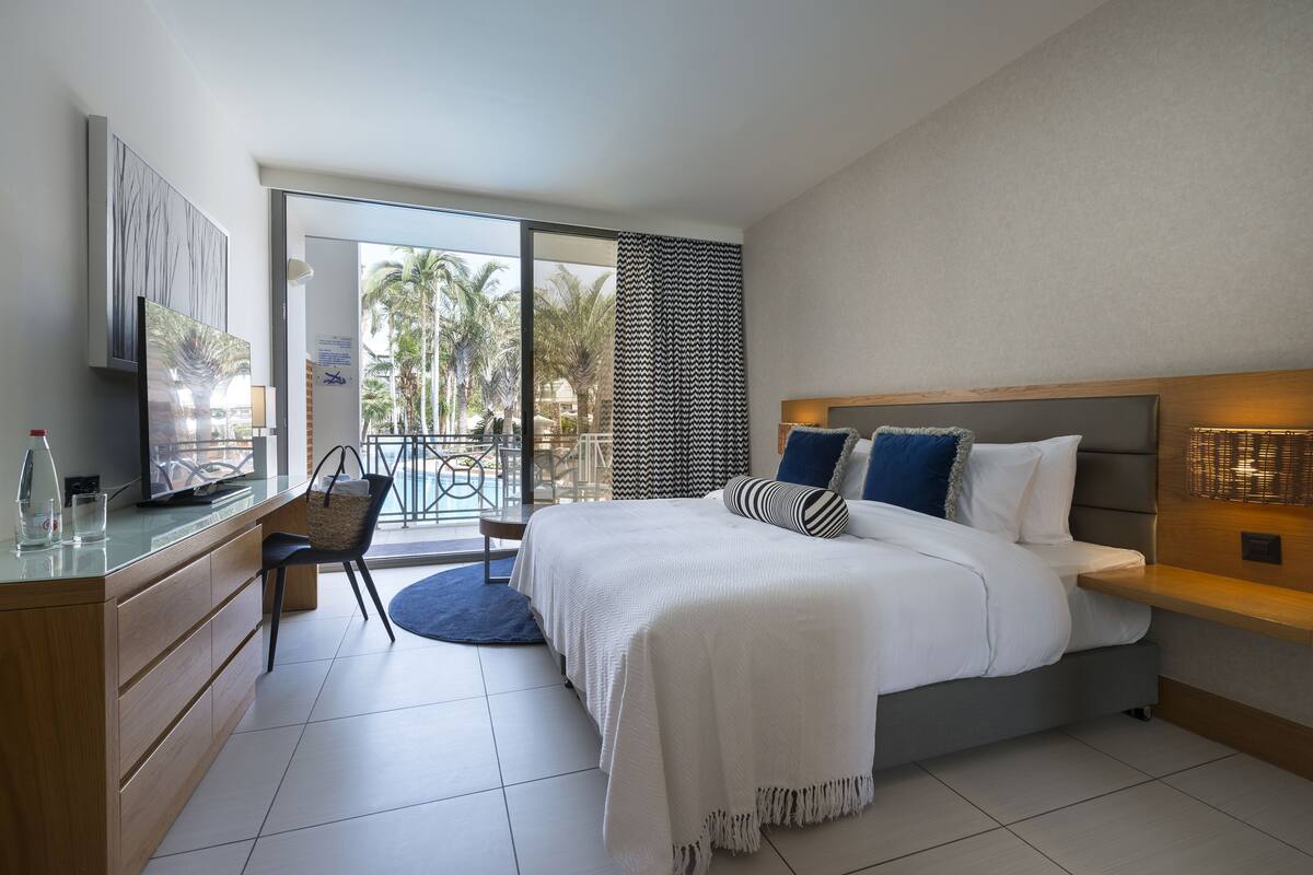 A modern hotel room with a large bed, a desk with a chair, a television, and a balcony with a view of palm trees.