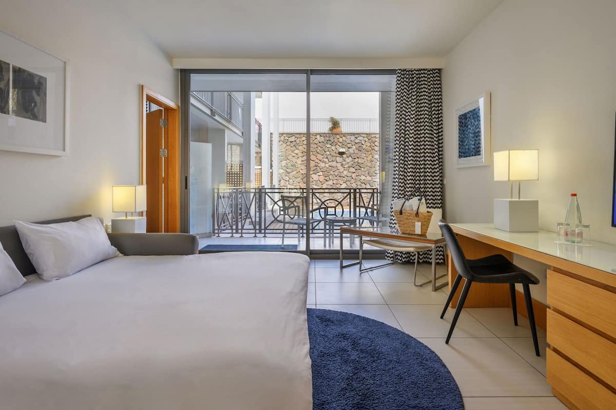 A hotel room with a bed, desk, chair, and a view of a balcony with outdoor seating.