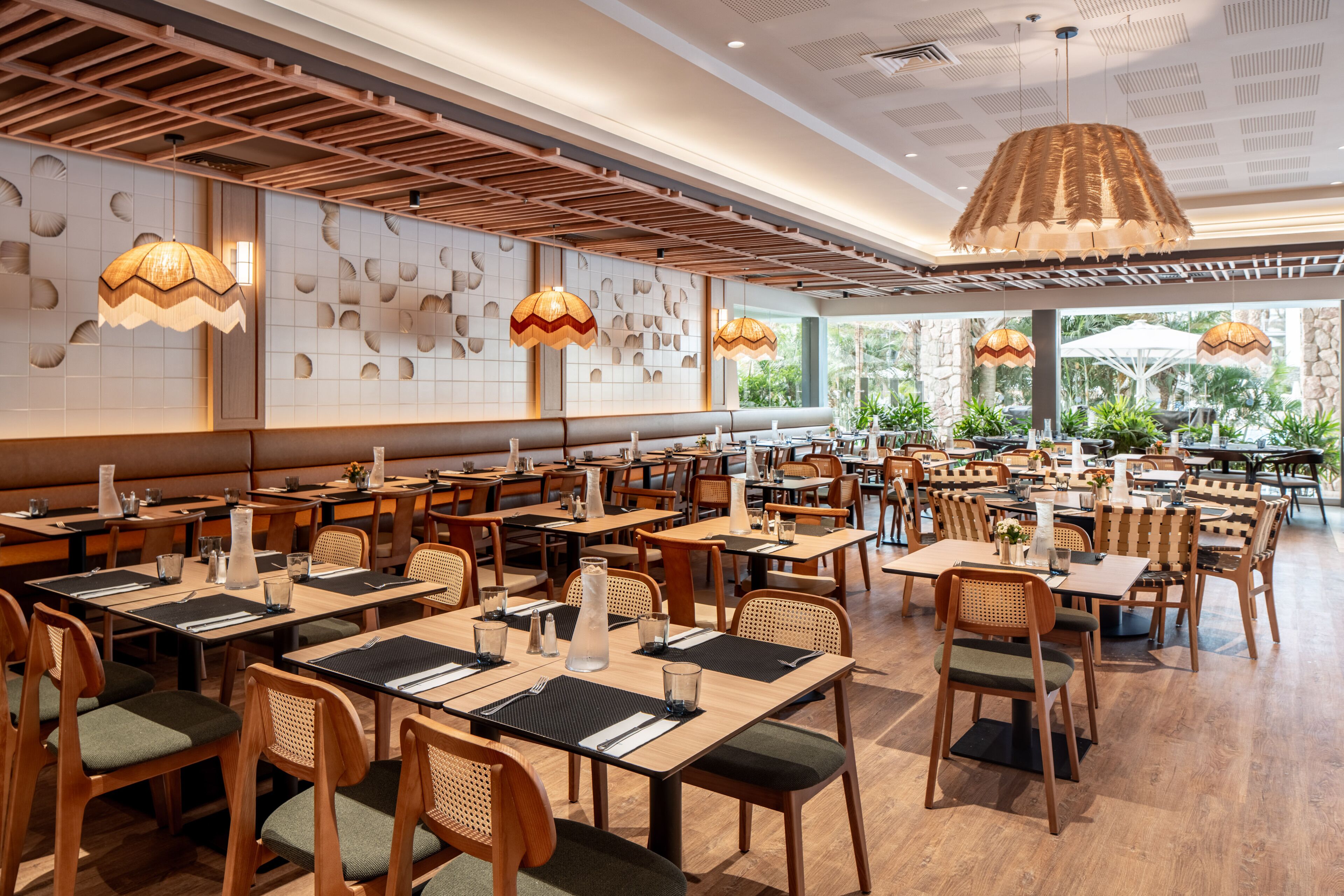 Un restaurante espacioso con varias mesas y sillas, techo de madera y grandes ventanales que dejan entrar luz natural.
