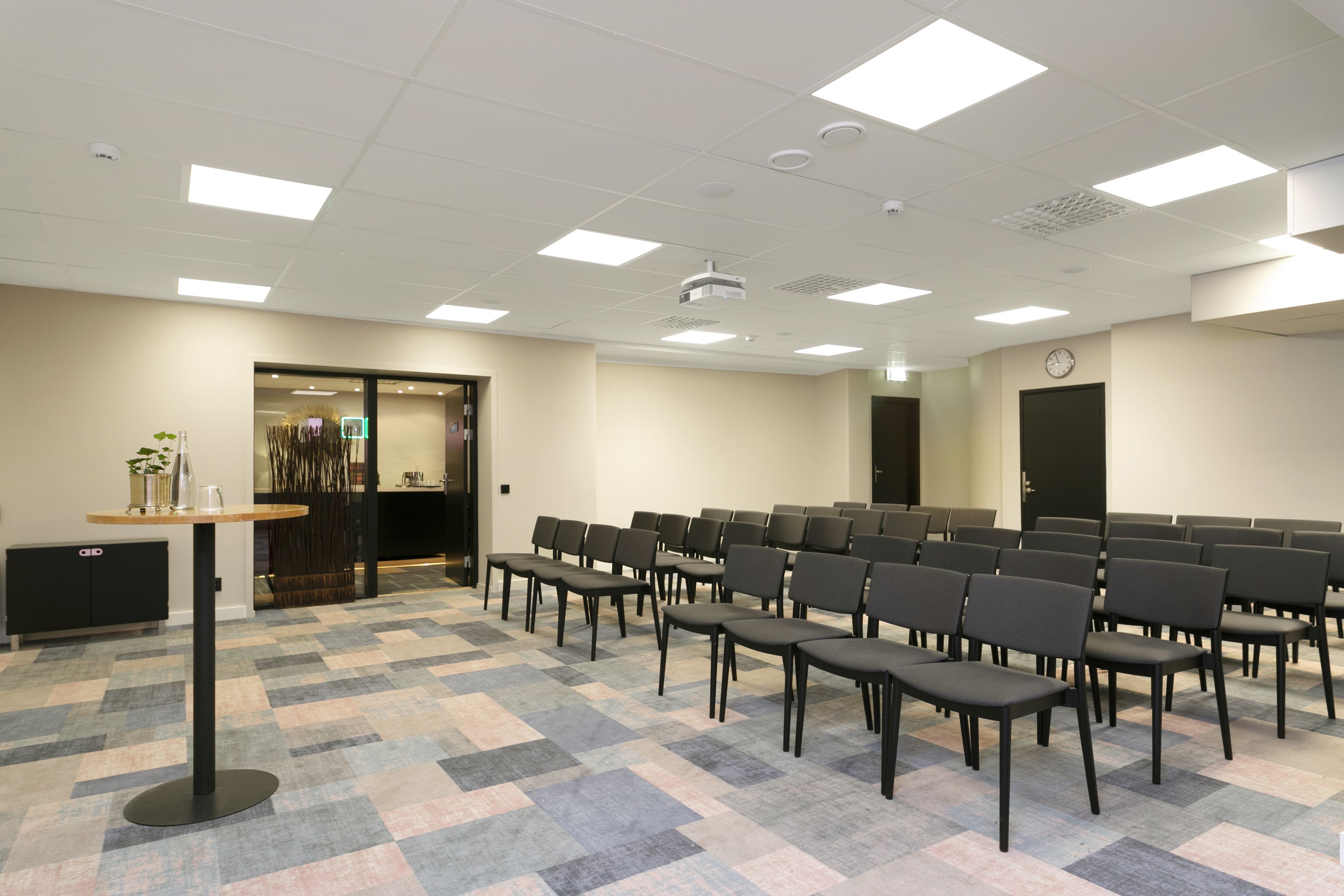 Un salón de conferencias con una larga mesa y filas de sillas, un reloj en la pared y una puerta que da a otra habitación.