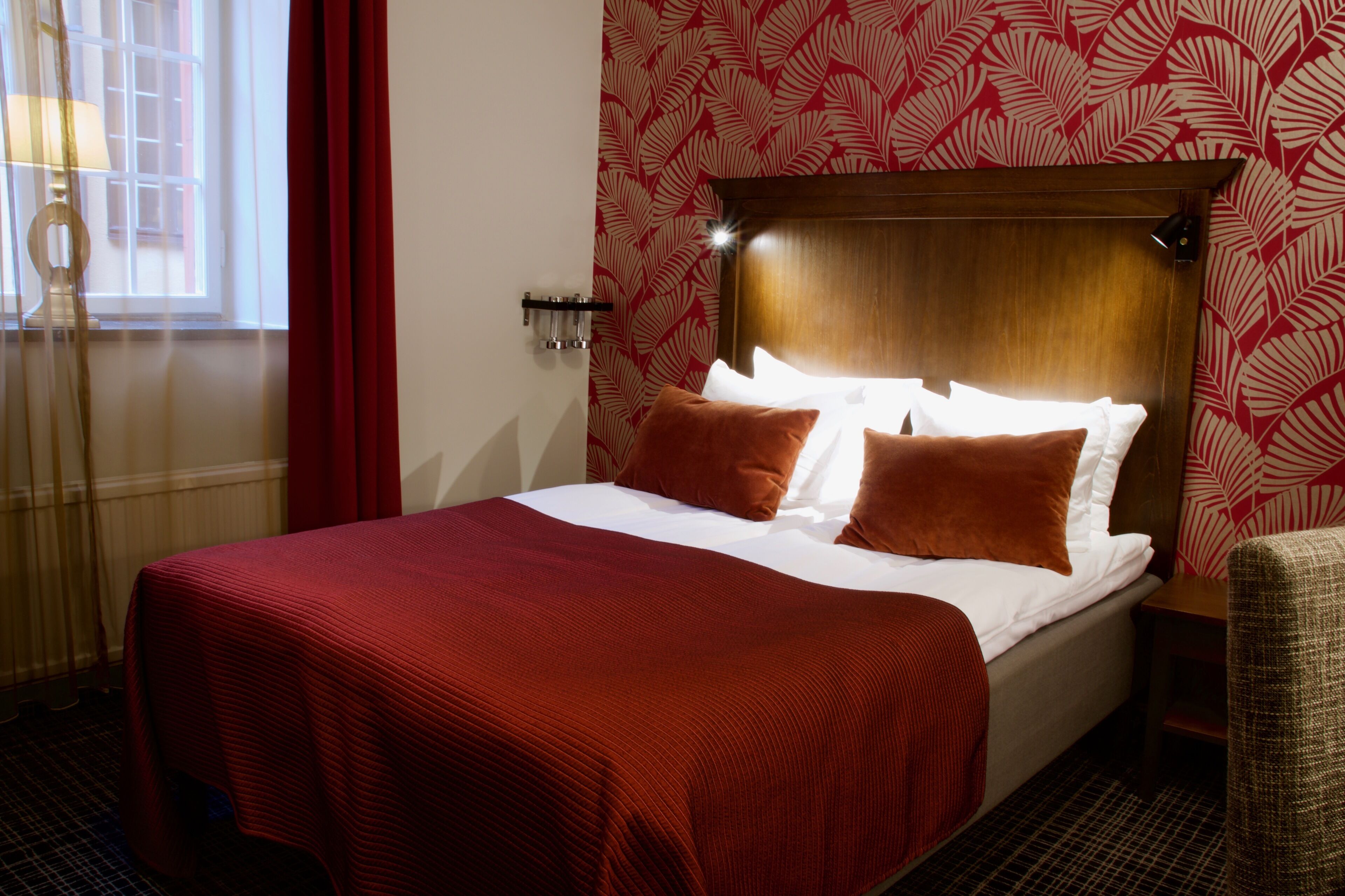 Ett hotellrum med en stor säng, röda gardiner, en mönstrad tapet och en sänggavel i trä.