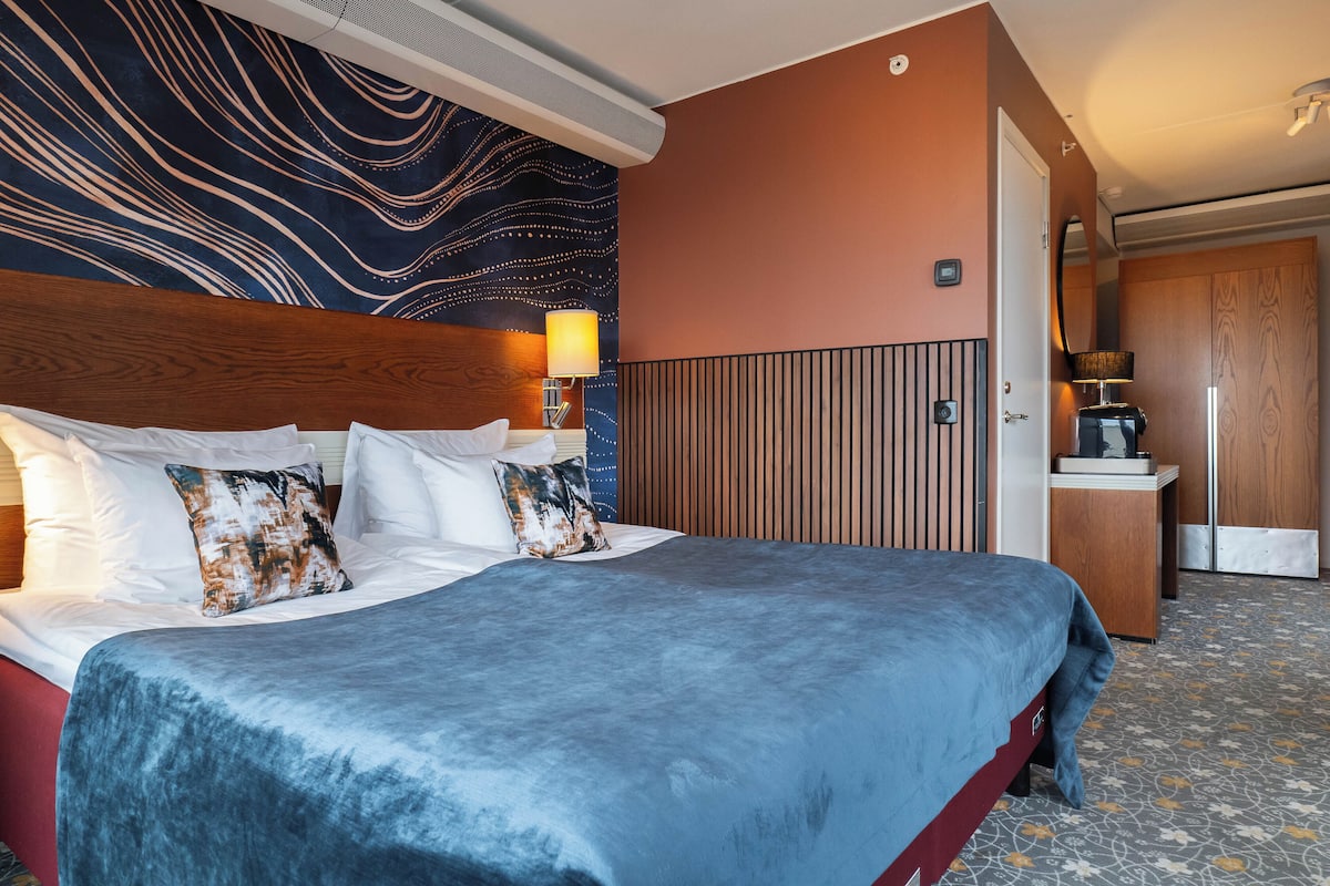 A hotel room with a large bed, a wooden headboard, a wall with a wavy pattern, a bedside table with a lamp, and a door leading to a bathroom.