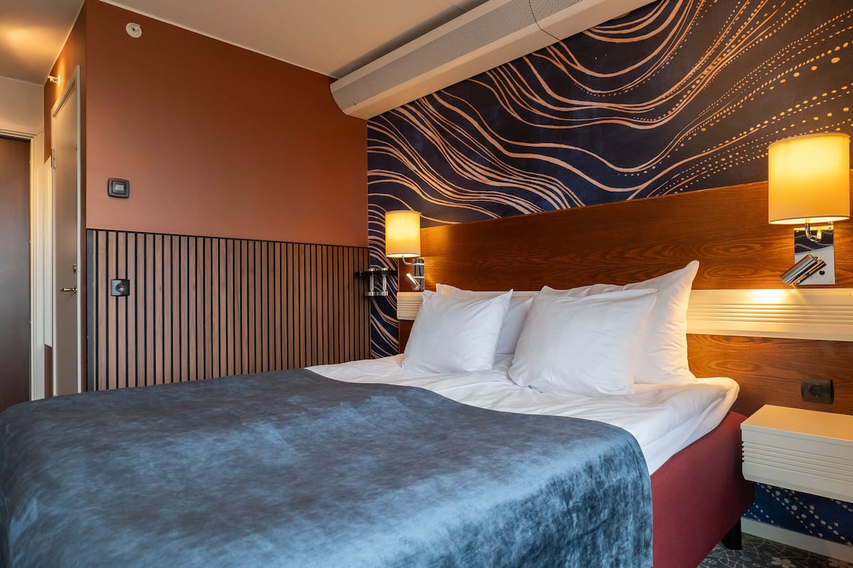 A hotel room with a large bed, a wooden headboard, two bedside lamps, and a patterned wall.