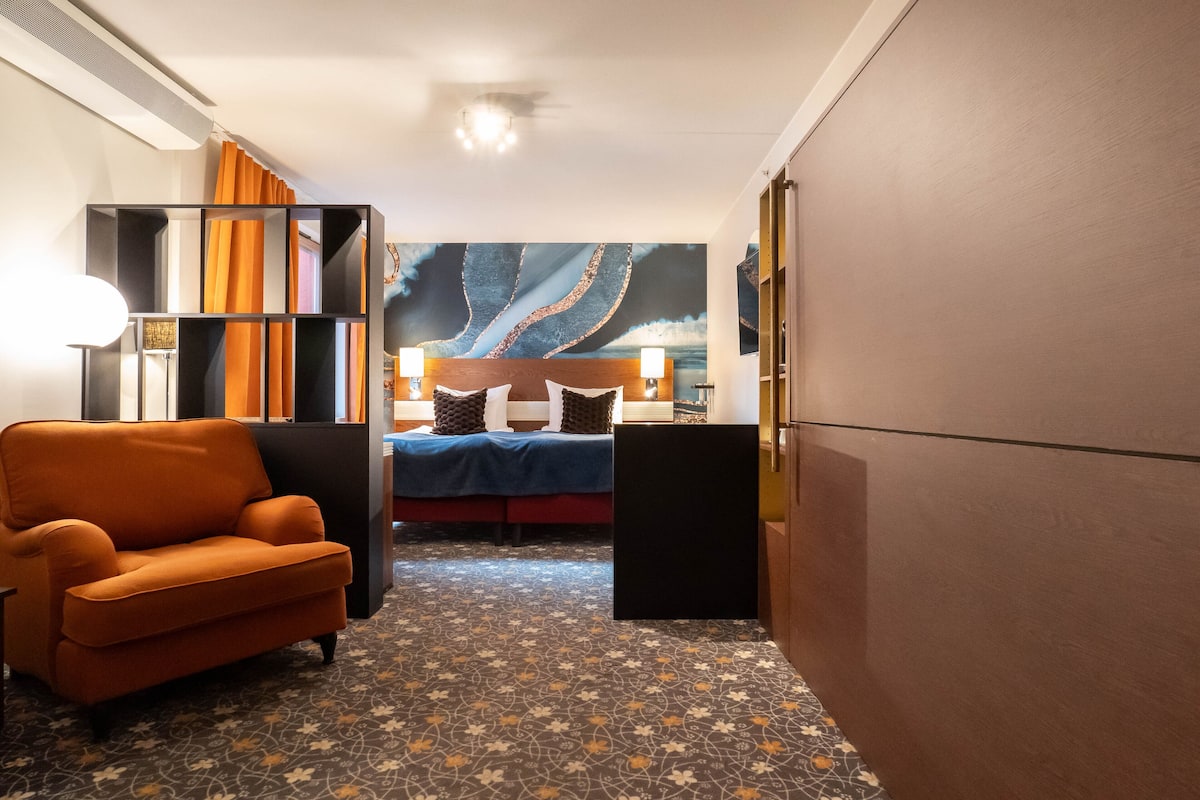 A hotel room with a bed, a sofa, a patterned carpet, and a large abstract painting on the wall.