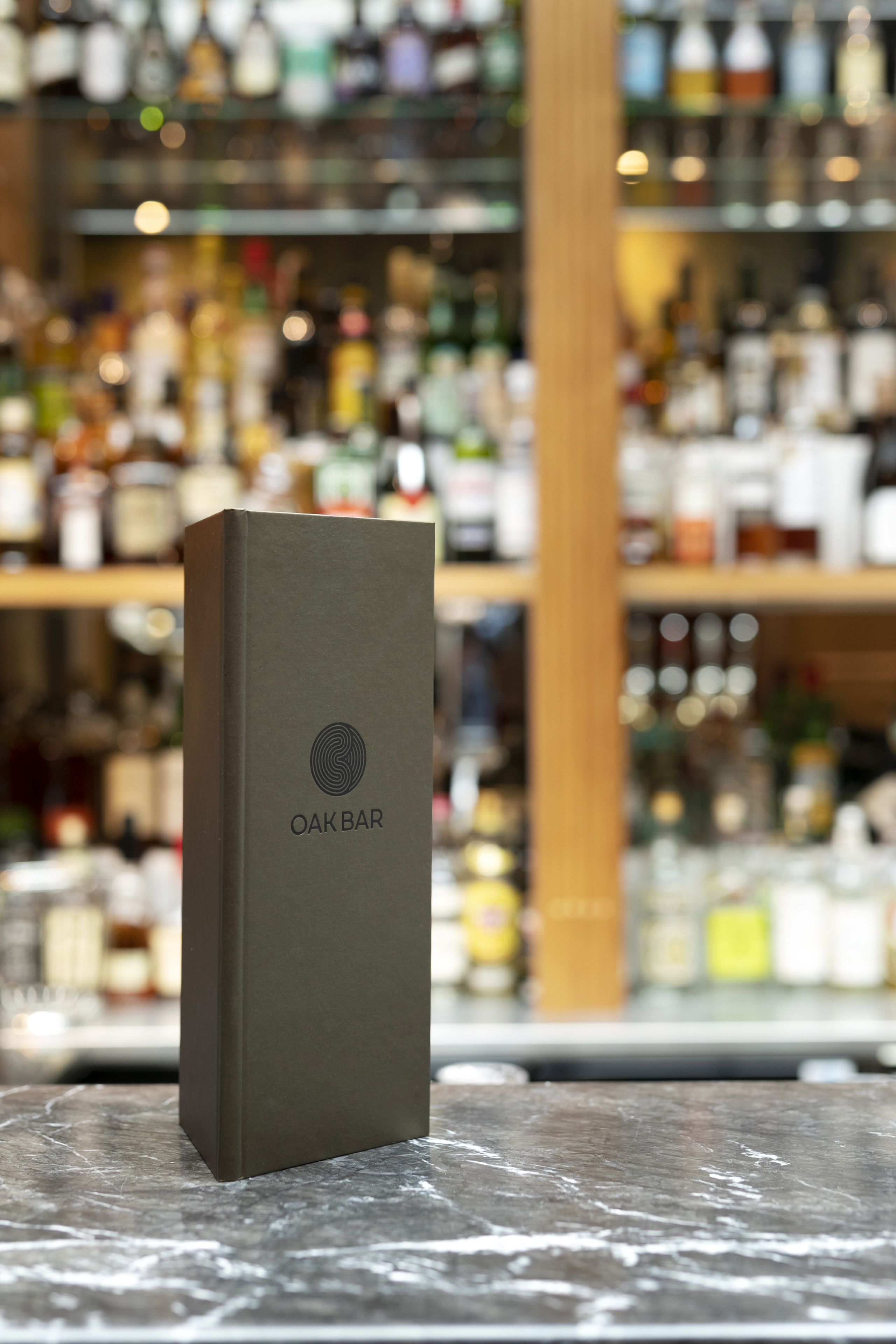 Una caja de color oscuro con el texto "OAKBAR" sobre ella, colocada sobre una superficie con un fondo desenfocado de varias botellas.