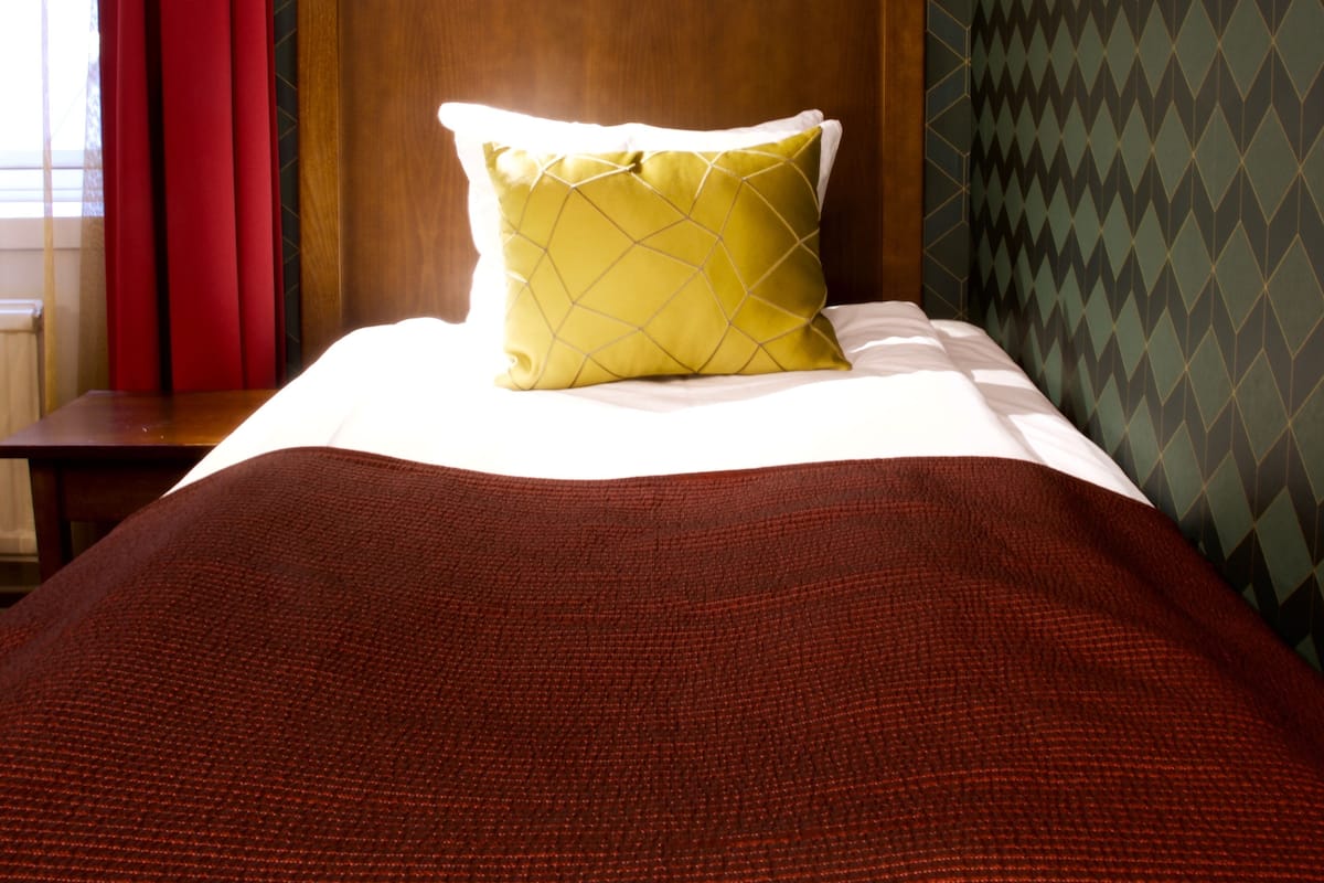 A neatly made bed with a red blanket and a yellow pillow in a room with patterned wallpaper and a wooden headboard.
