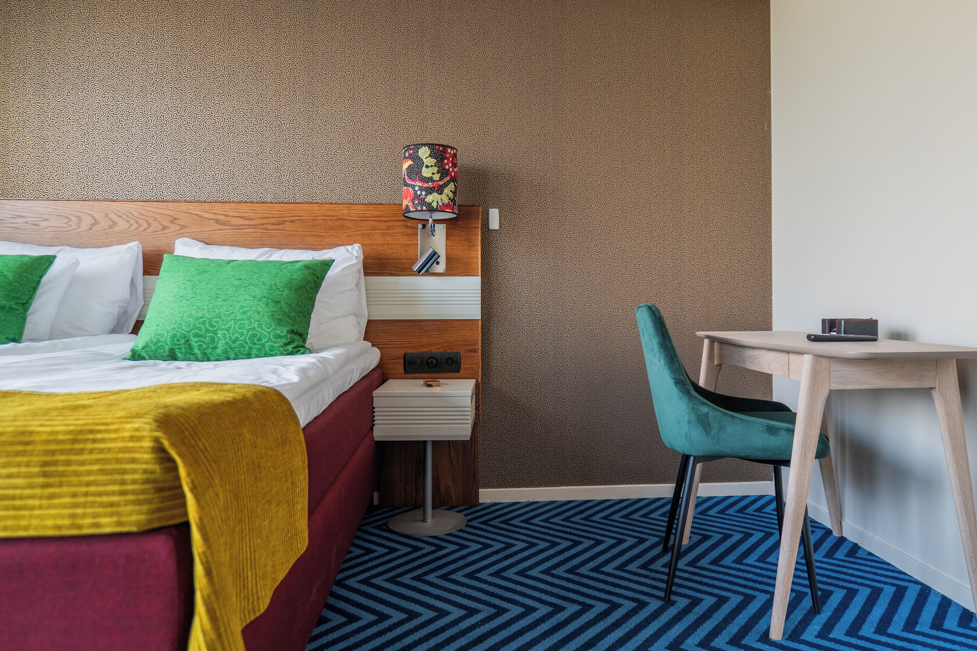 A hotel room with a bed, bedside table, lamp, chair, and a desk.