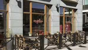 Un espace extérieur aménagé avec des chaises et des tables en osier, une structure vitrée sur laquelle on peut lire le nom « Angelini », et une façade en pierre.