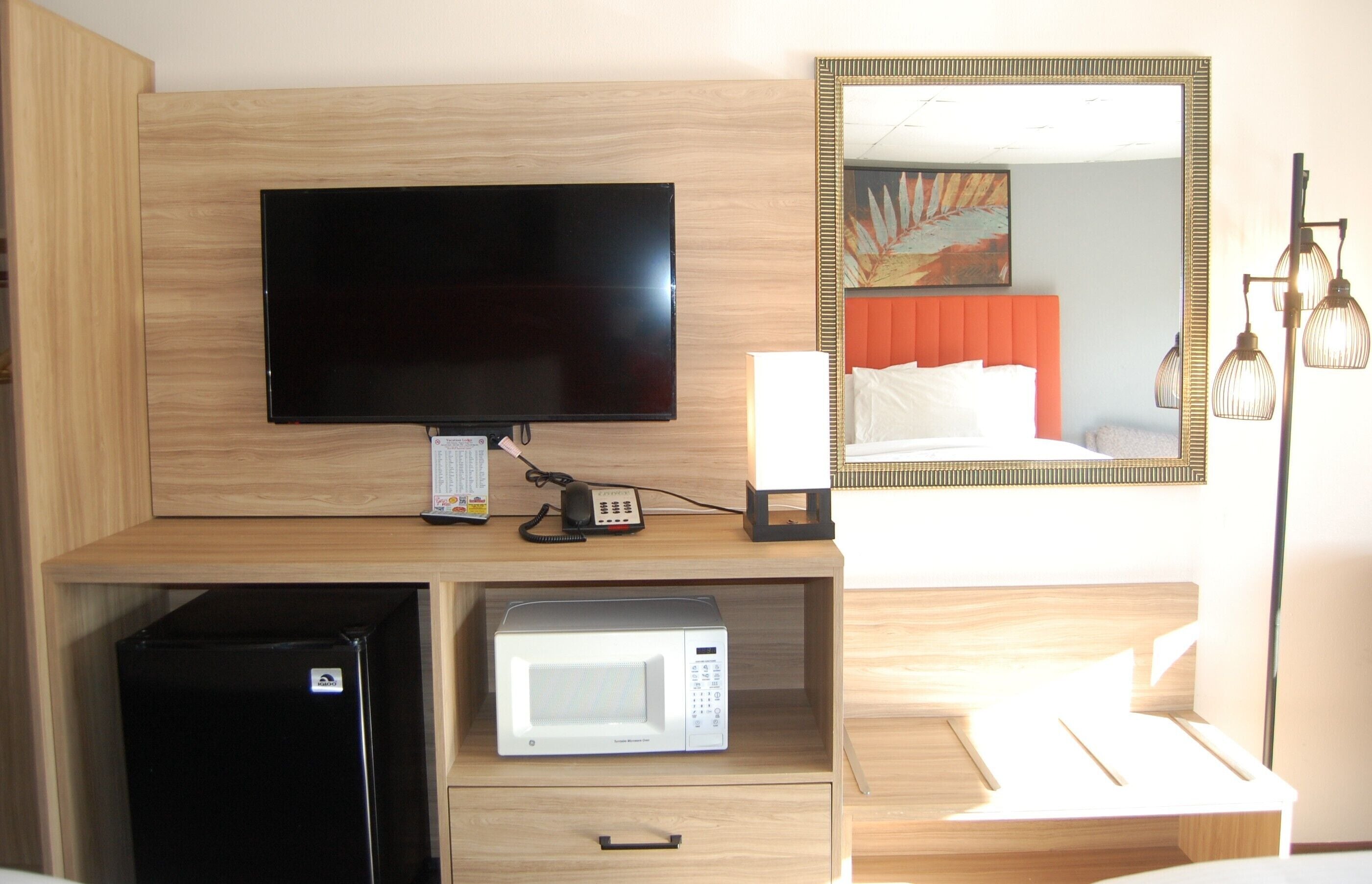 A hotel room with a television, a microwave, a refrigerator, a bedside table, a lamp, and a bed with an orange headboard.