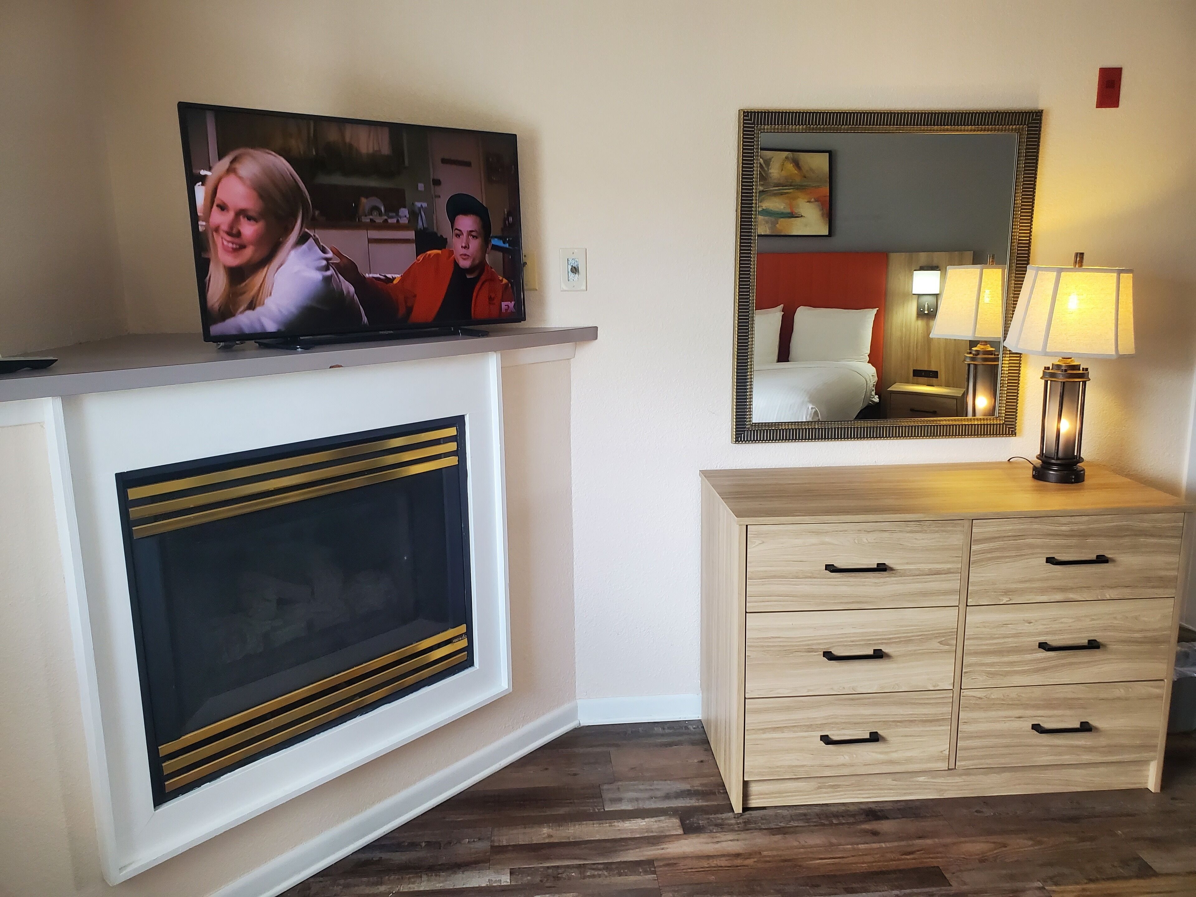Une pièce avec une cheminée, un téléviseur fixé au-dessus, un miroir et une table de chevet avec une lampe.