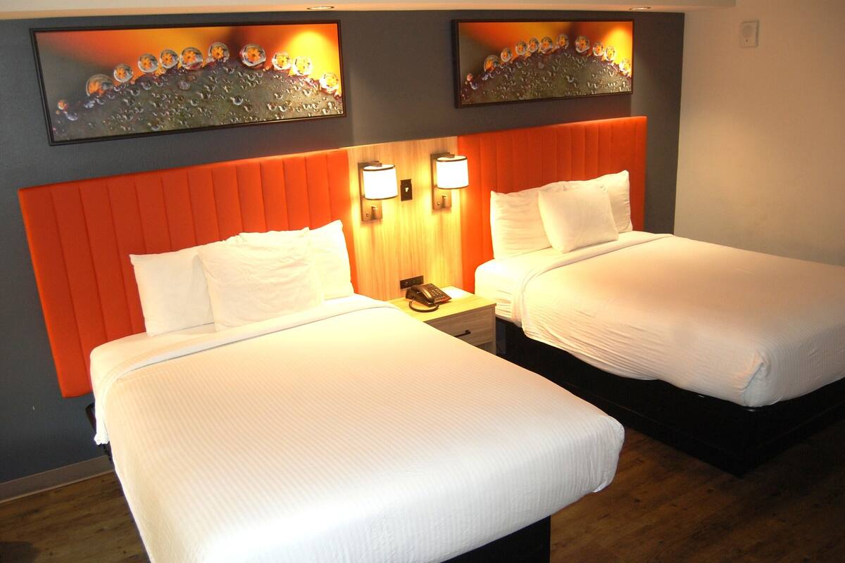 A hotel room with two beds, each with white linens, a wooden nightstand with a lamp, and a wall-mounted artwork above the beds.