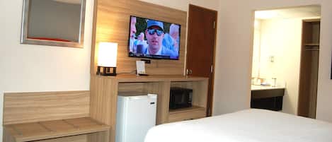 A hotel room with a bed, a television mounted on a wooden cabinet, a small refrigerator, and a wooden bench.