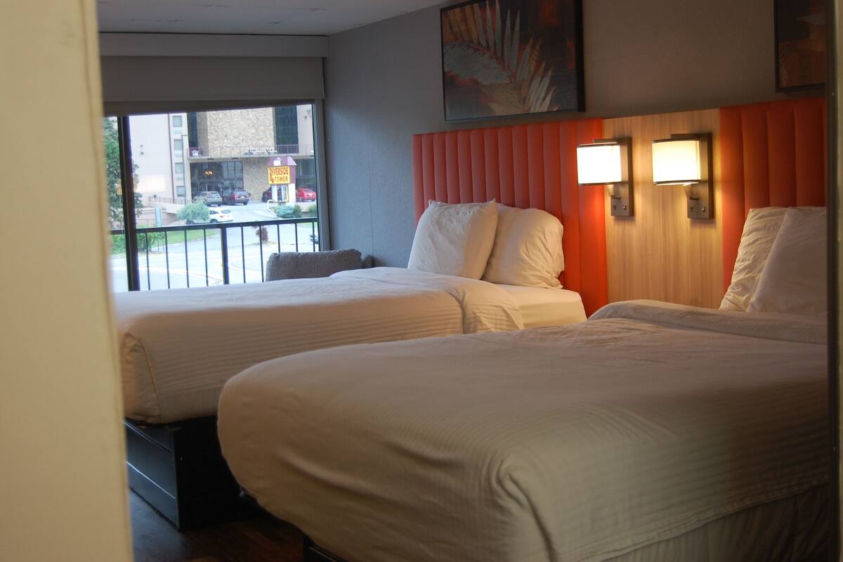A hotel room with two beds, a window with a view of the street, and a wooden headboard with wall-mounted lights.