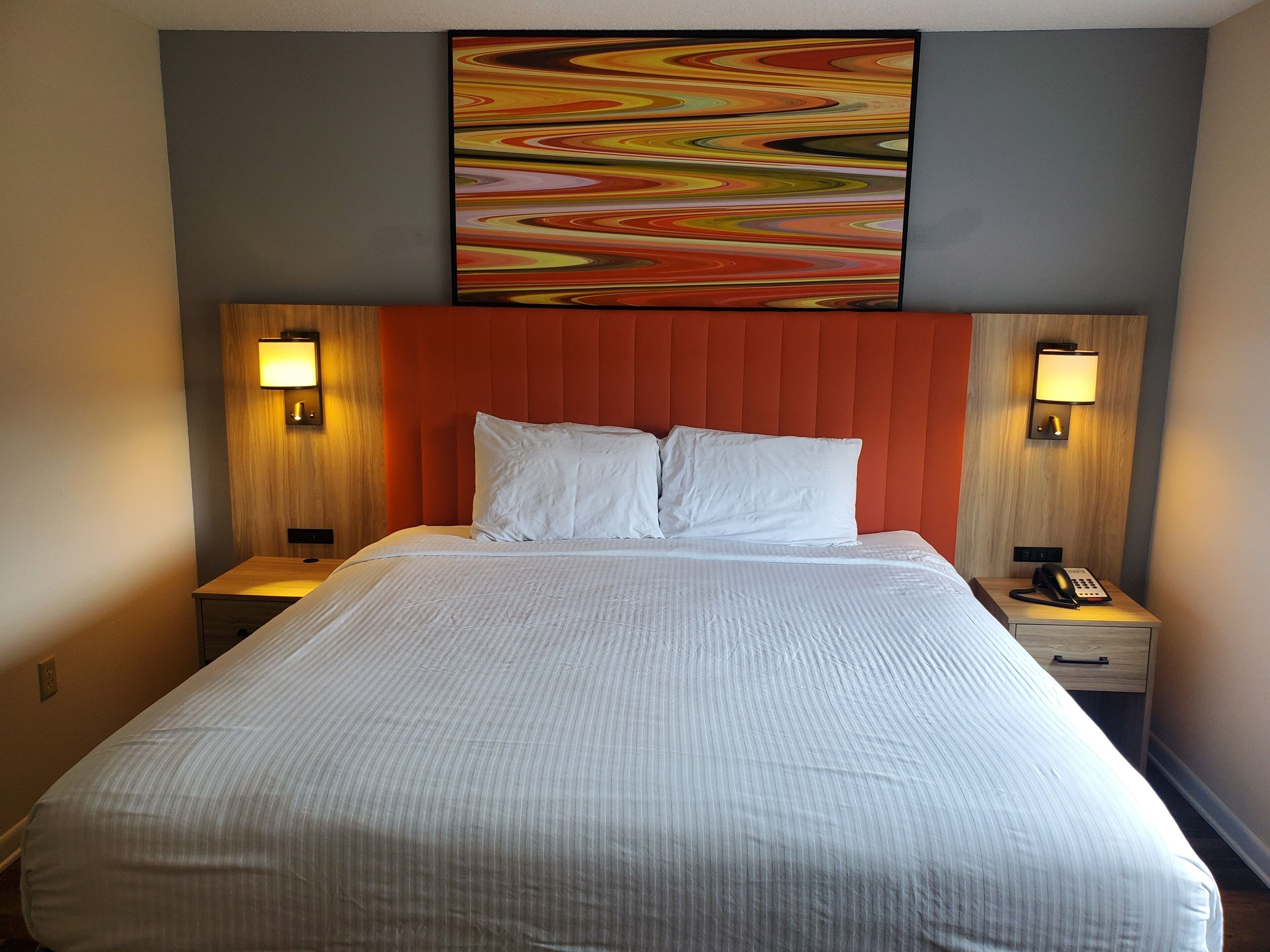 Hotelkamer met een groot bed, twee nachtkastjes met lampen, een rood hoofdeinde en een kleurrijk abstract schilderij boven het bed.