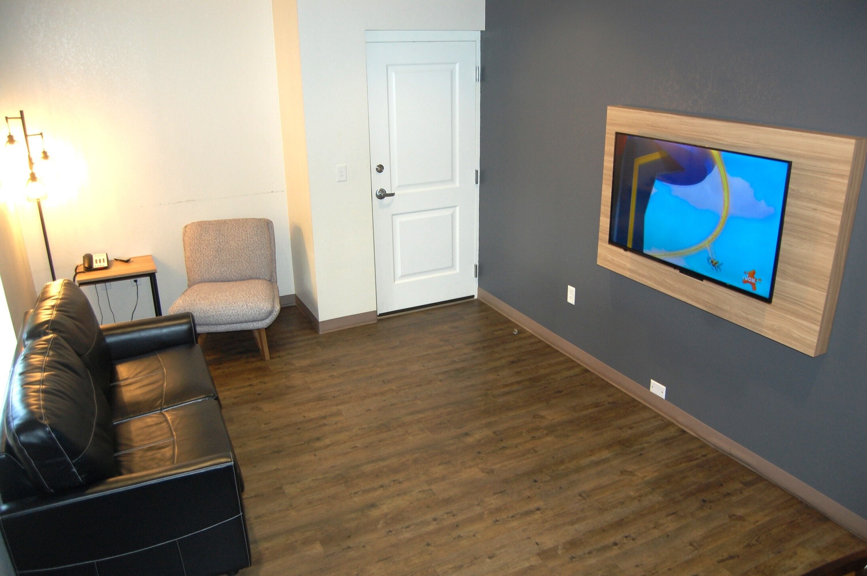 Una sala de estar con un sofá de cuero negro, un sillón gris, un mueble bajo para televisión de madera con un televisor montado, una puerta blanca y una lámpara sobre una mesita.