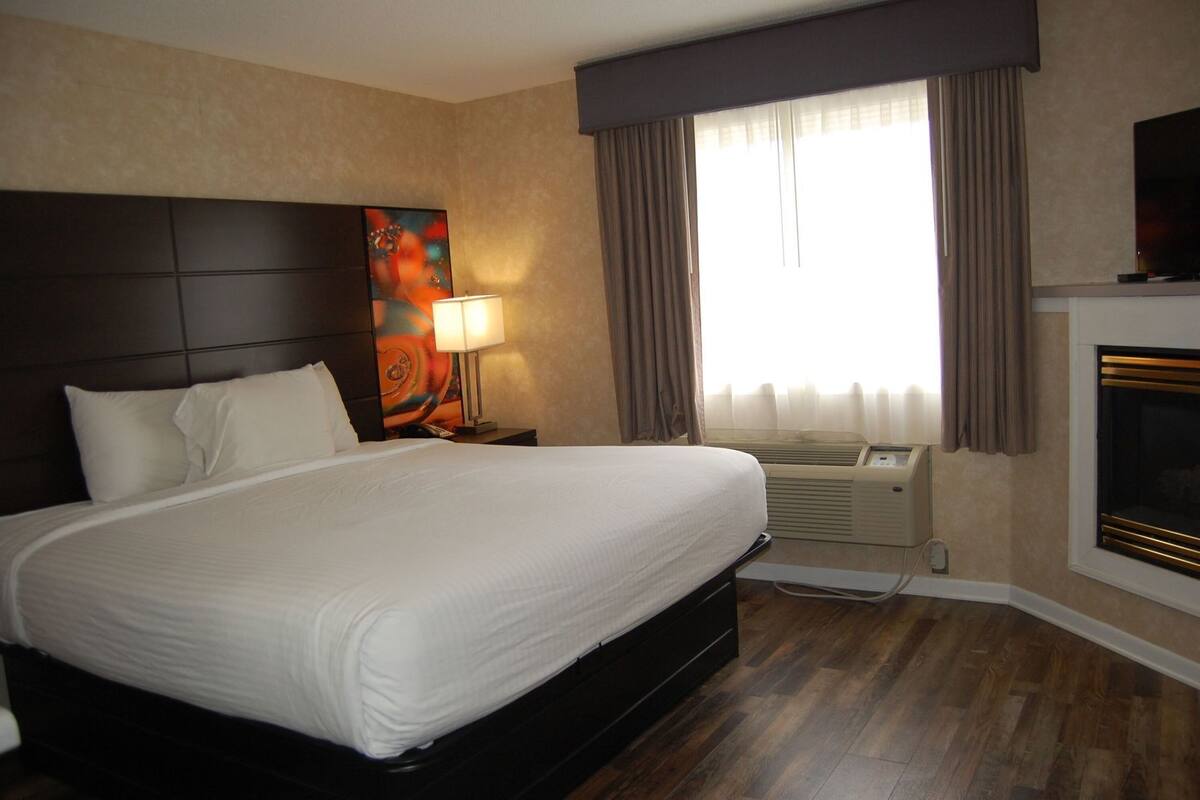 A hotel room with a large bed, a painting on the wall, a window with curtains, and a fireplace.