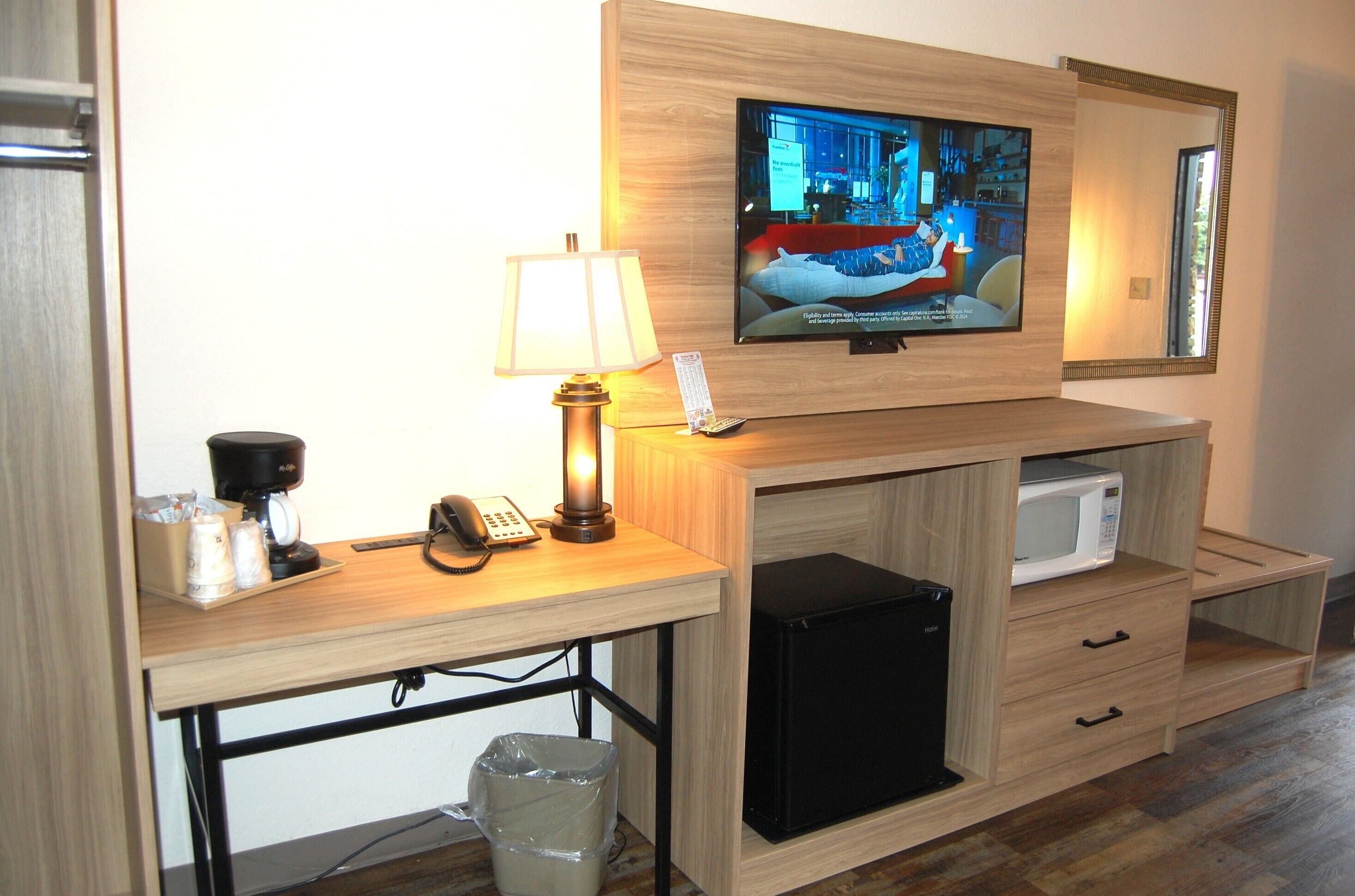 Een hotelkamer met een aan de muur bevestigde televisie, een bureau met een lamp, een koffiezetapparaat, een telefoon, een magnetron en een prullenbak.