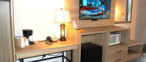 Een hotelkamer met een aan de muur bevestigde televisie, een bureau met een lamp, een koffiezetapparaat, een telefoon, een magnetron en een prullenbak.