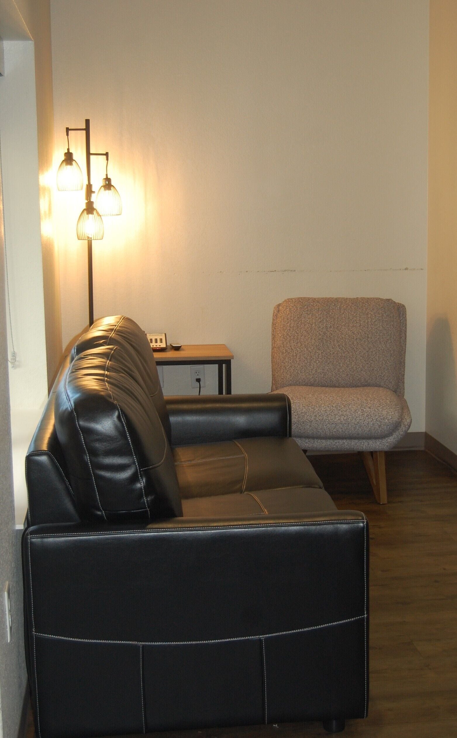 A black leather sofa and a grey armchair in a room with a wooden floor and a lamp with three shades.