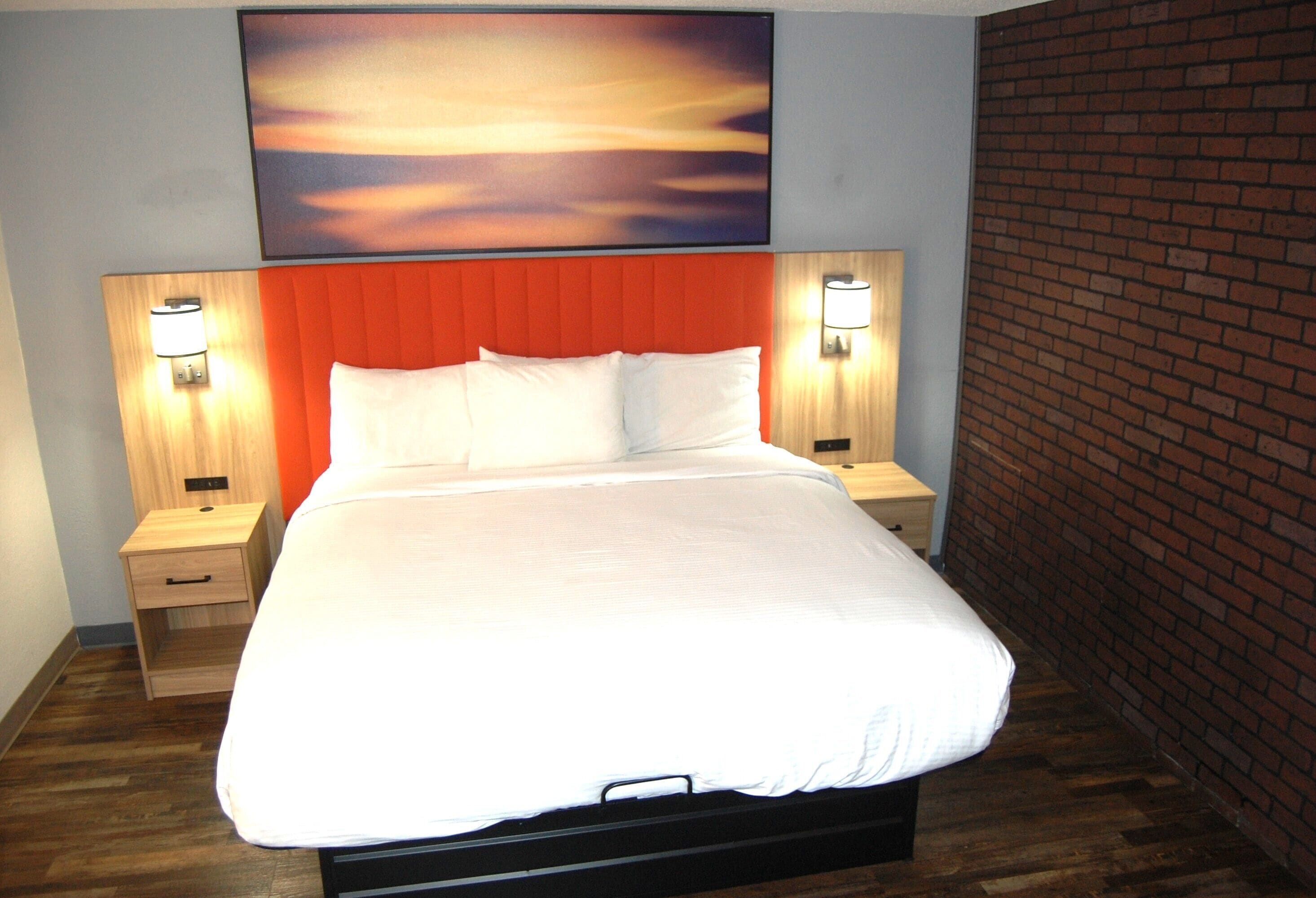 Habitación de hotel con una cama grande, dos mesitas de noche con lámparas, un cabecero de madera, una pared de ladrillo y un cuadro de atardecer sobre la cama.