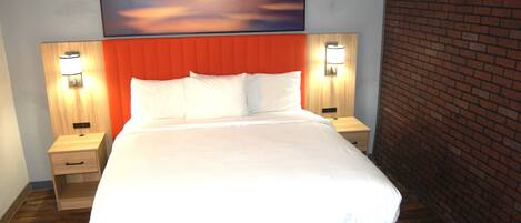Habitación de hotel con una cama grande, dos mesitas de noche con lámparas, un cabecero de madera, una pared de ladrillo y un cuadro de atardecer sobre la cama.