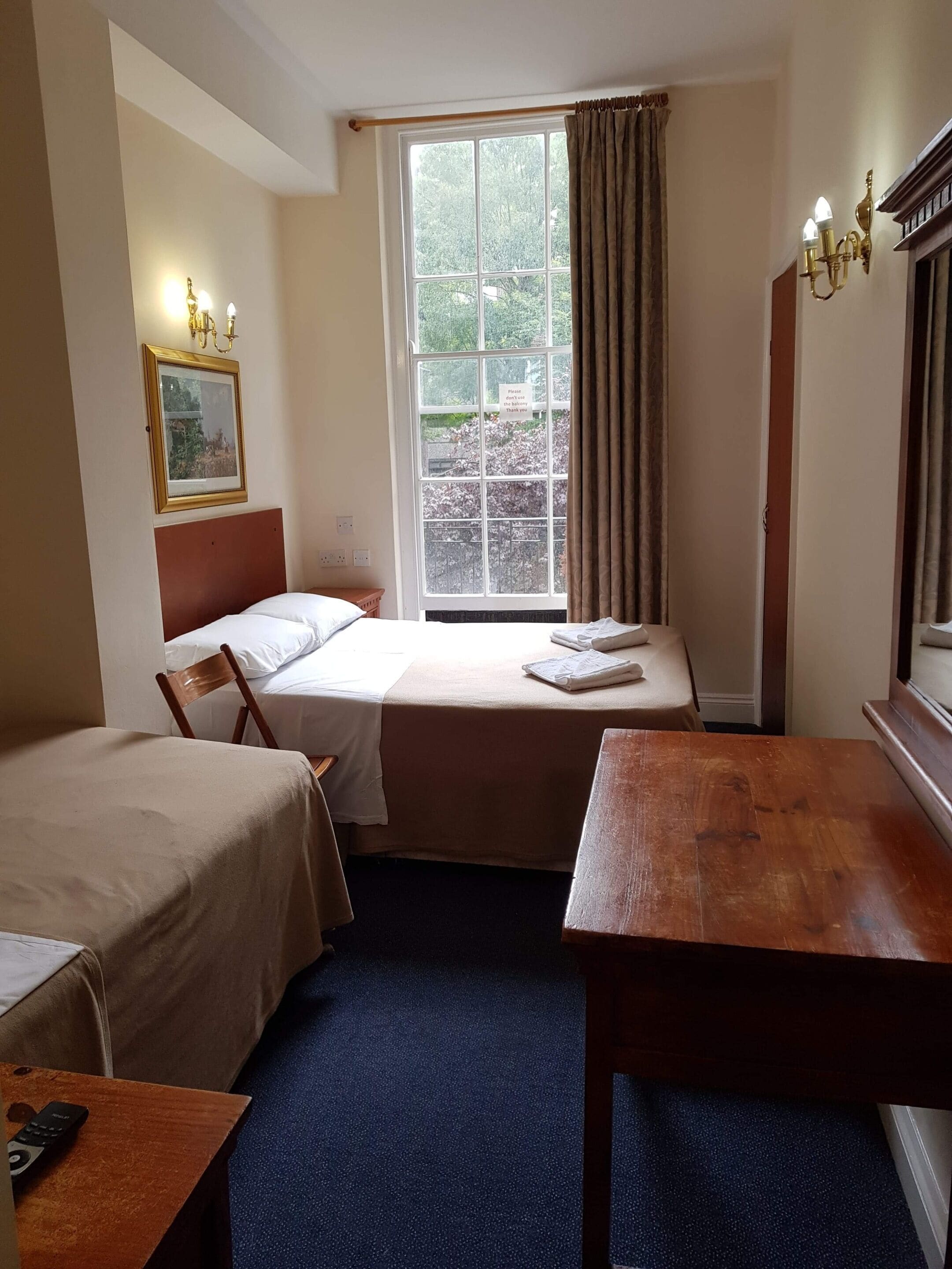 A hotel room with two beds, a wooden desk, a window with curtains, a framed picture on the wall, and a sign thanking guests.