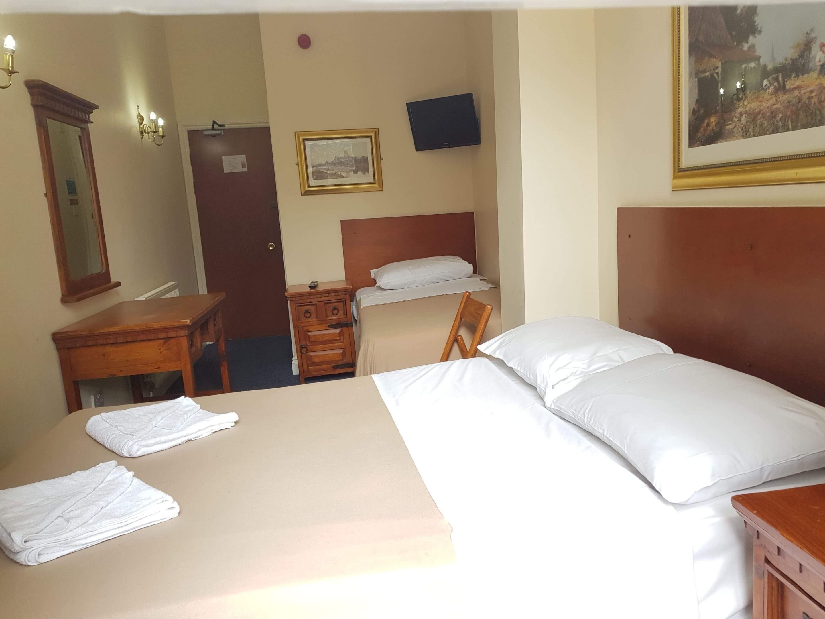 Habitación de hotel con cama, mesita de noche de madera, una silla, escritorio, espejo, cuadro enmarcado y televisor.