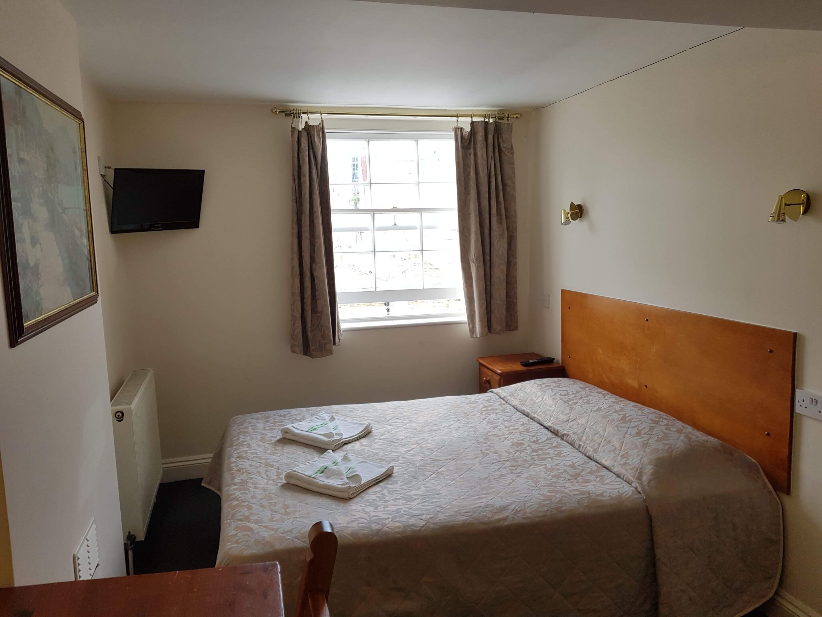 Ein Hotelzimmer mit einem Einzelbett, einem Nachttisch, einem an der Wand befestigten Fernseher und einem Fenster mit Vorhängen.