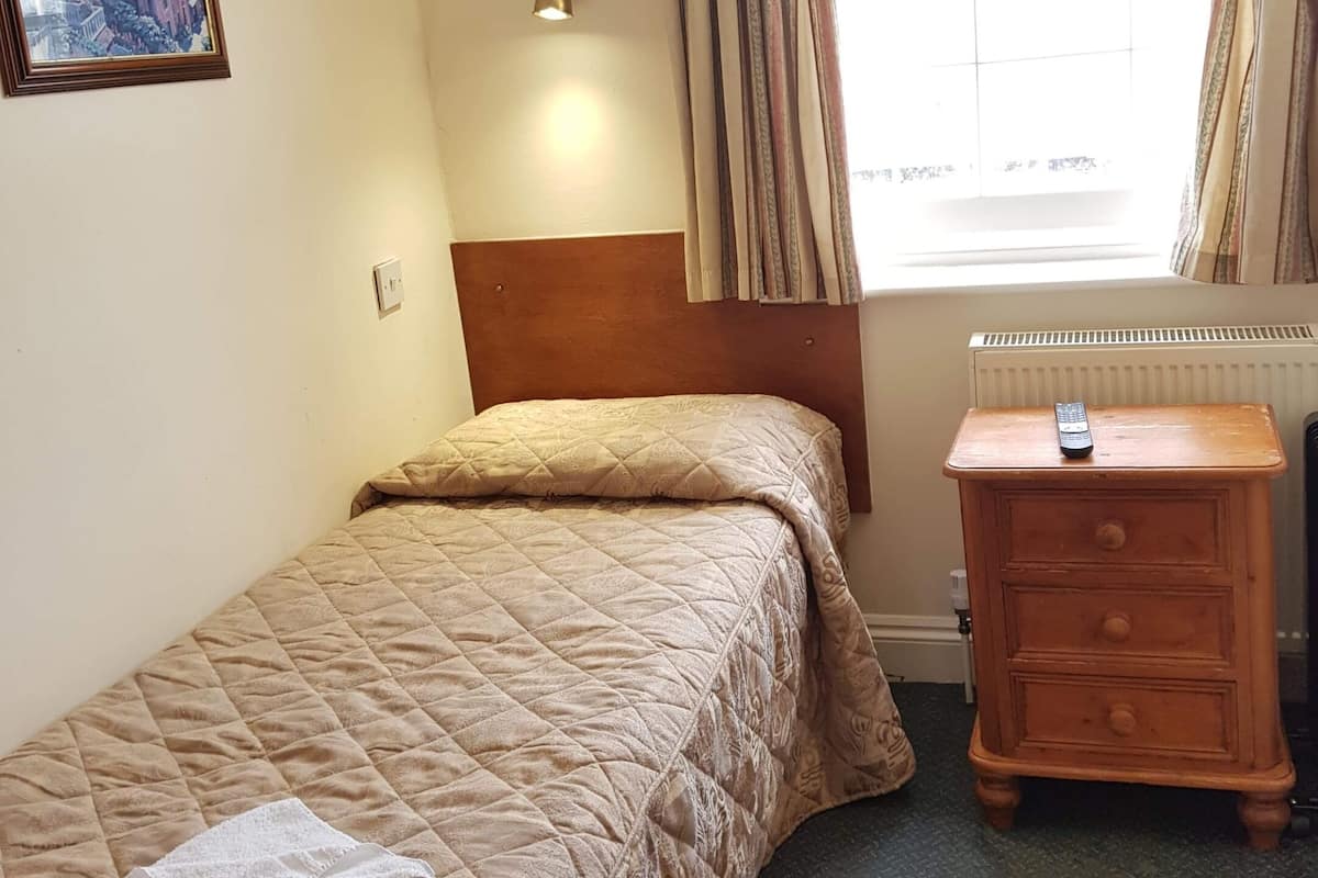 A bedroom with a bed, a nightstand, a lamp, a window with curtains, and a painting on the wall.