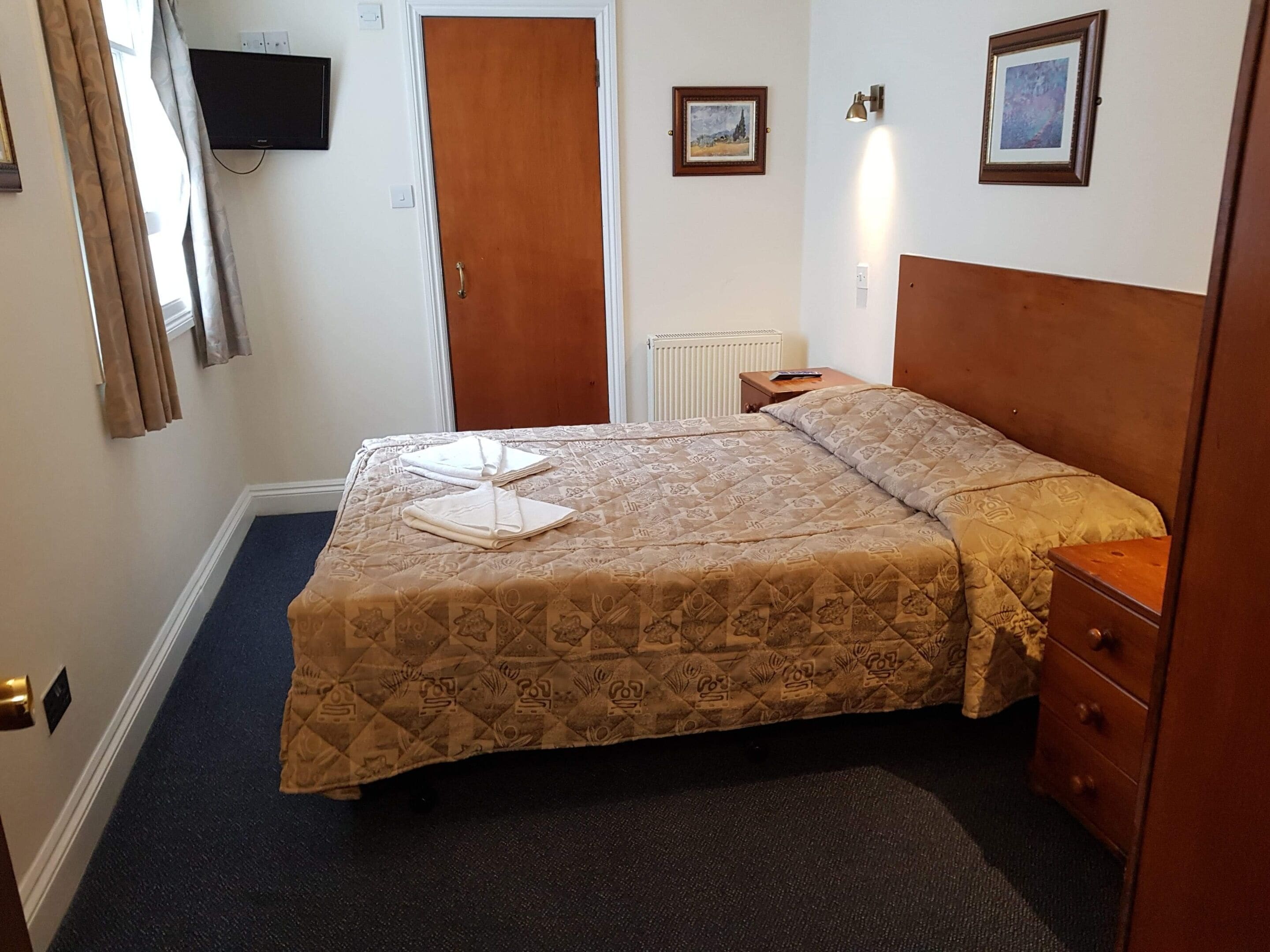 A neatly arranged hotel room with a bed, a nightstand, a dresser, a television mounted on the wall, and a door.