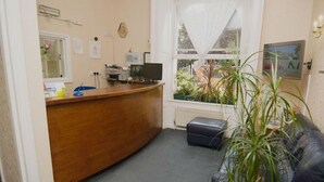 A reception area with a wooden desk, a computer, and a window with a view of the outside. - Normandie Hotel (London)