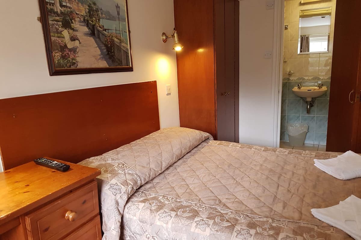A hotel room with a bed, a wooden nightstand, a remote control, a painting on the wall, and a bathroom visible through an open door.