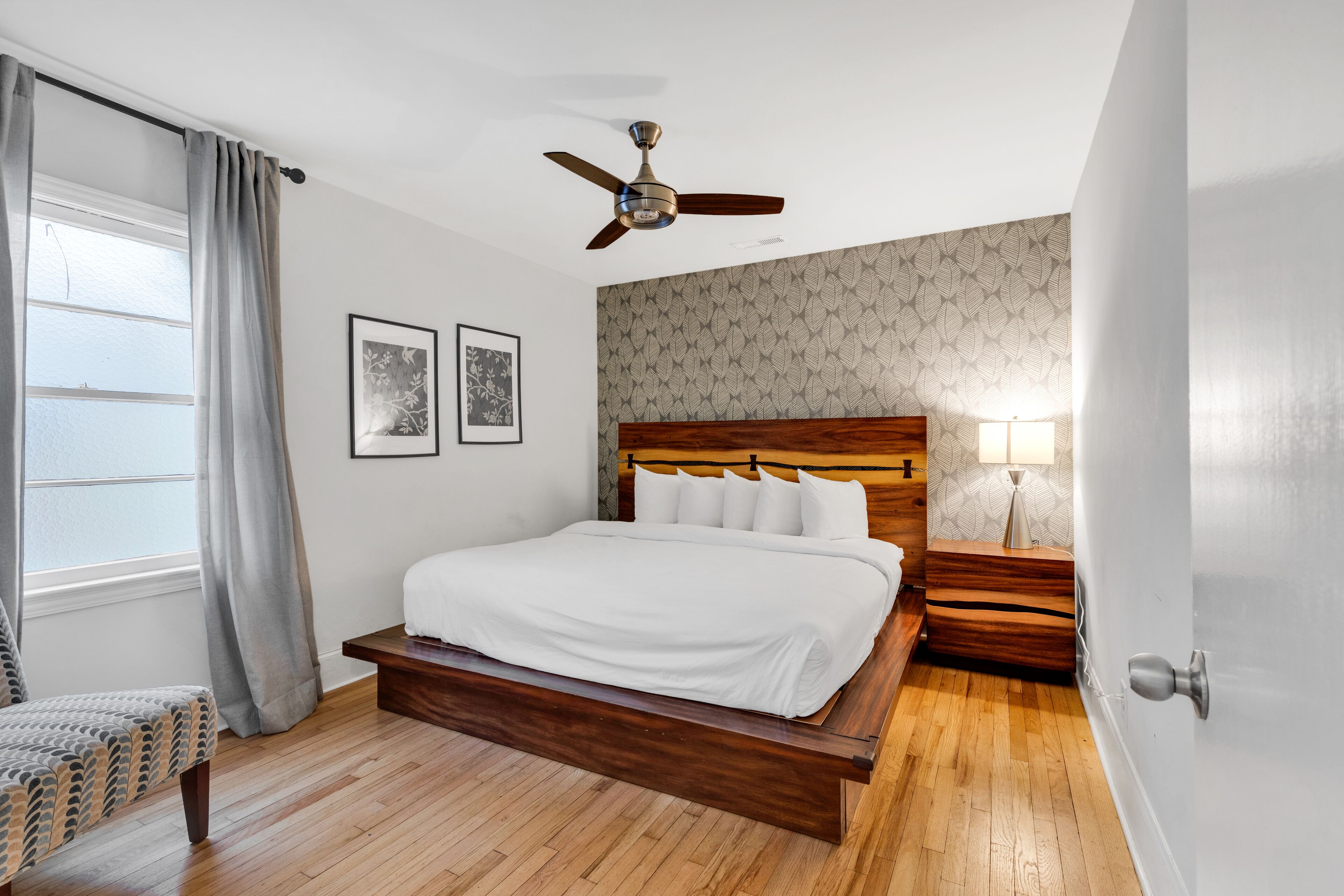 A bedroom with a wooden bed, a ceiling fan, a nightstand with a lamp, and a window with curtains.