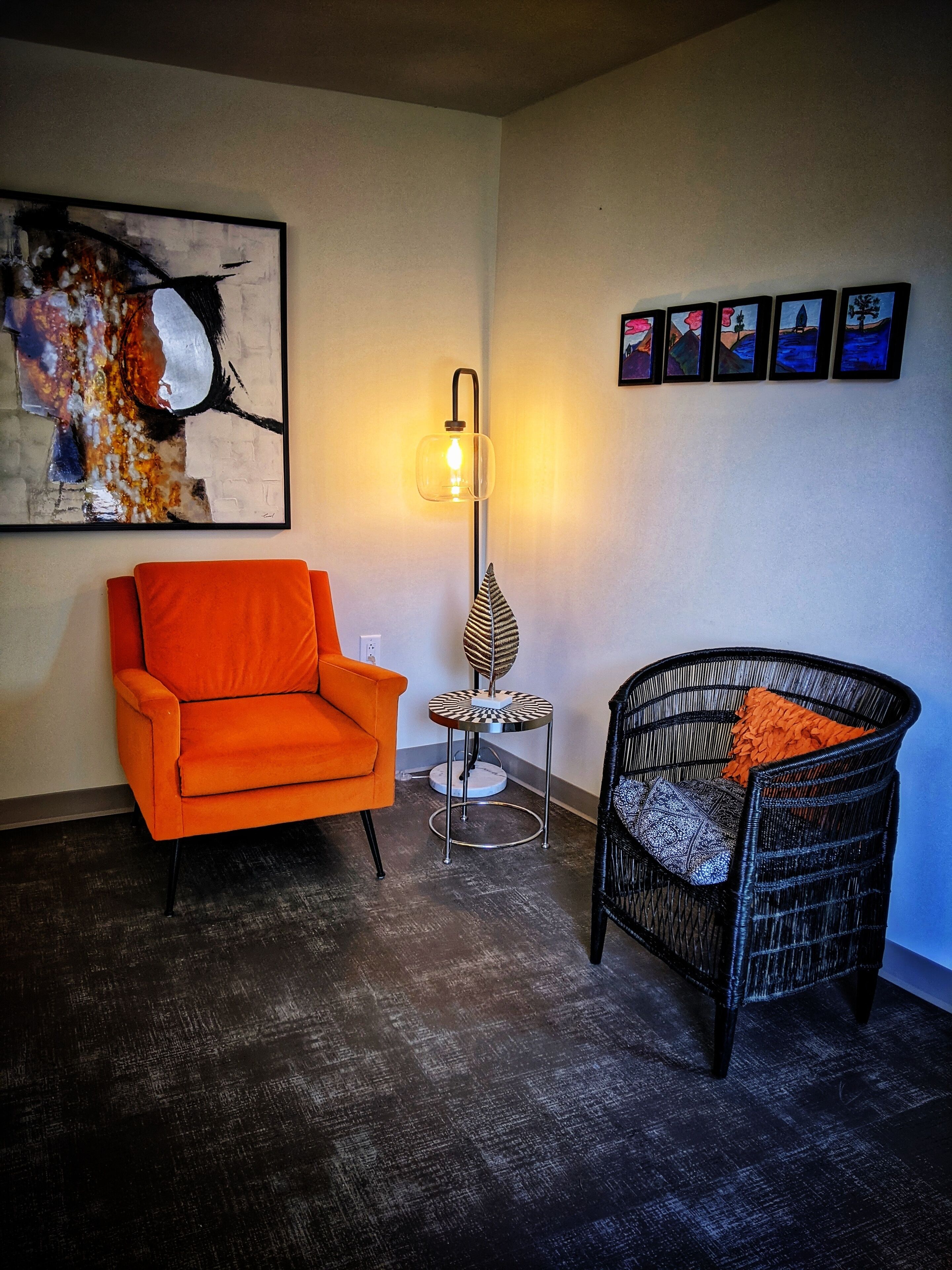 Una habitación con un sillón moderno color naranja, una butaca de mimbre negro con cojín naranja, una lámpara de pie y un cuadro abstracto grande en la pared.