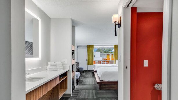 A hotel room with a large mirror, a white sink, a wooden vanity, a bed with white bedding, and a red door. - Holiday Shores (Myrtle Beach)