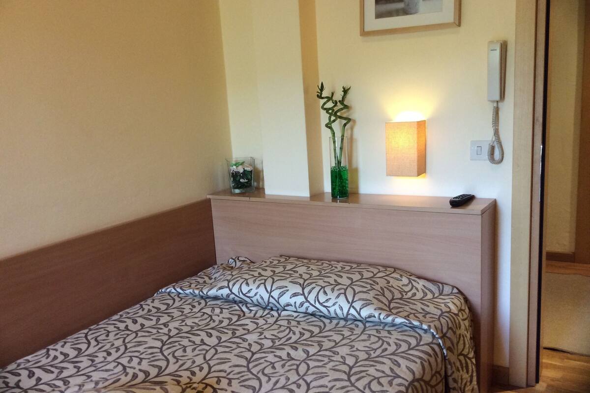 A neatly made bed with a patterned bedspread, a bedside table with a vase and a lamp, and a phone on the wall.