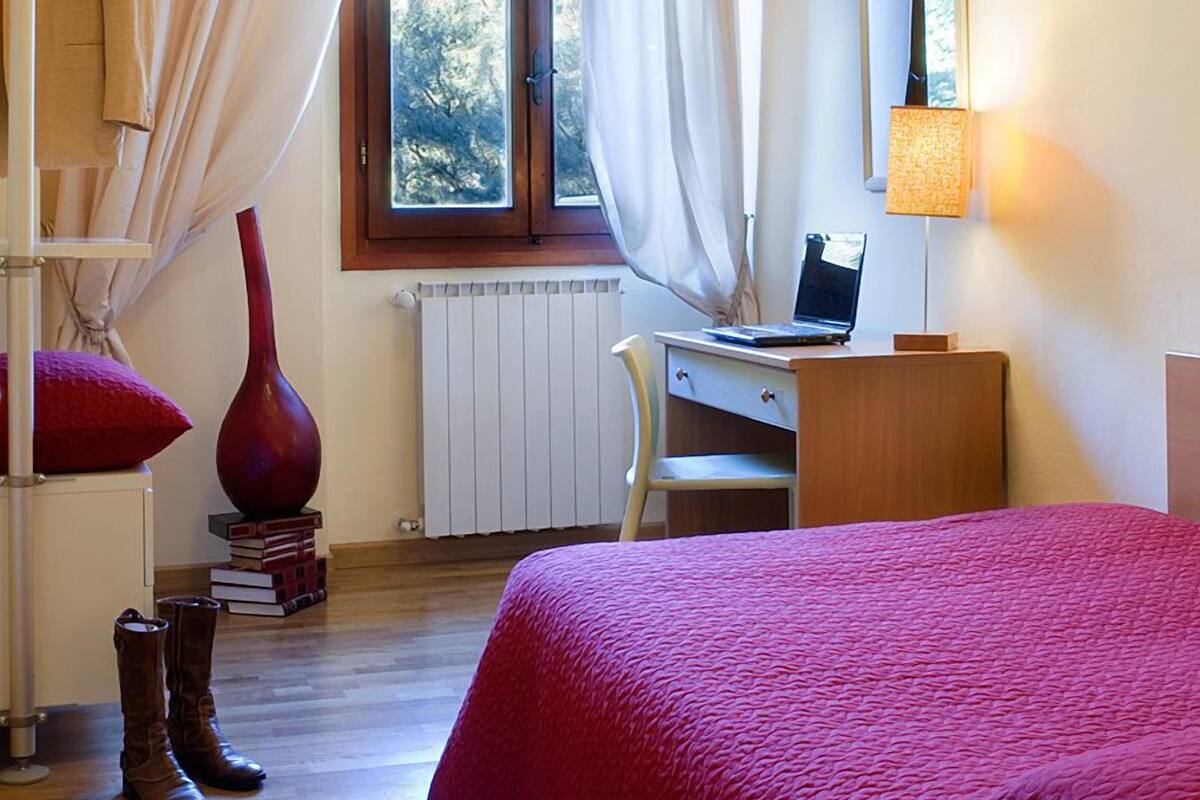 A bedroom with a bed, a desk, a chair, a lamp, a window with curtains, and a radiator.