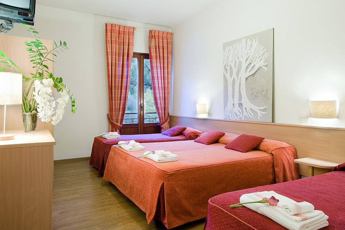 A hotel room with two beds, a desk, a window with curtains, a vase with flowers, and a wall art piece.