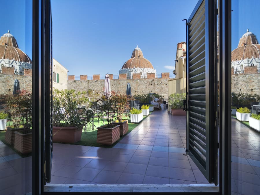 Pátio com piso de azulejos, plantas em vasos e vista para um edifício histórico com cúpulas.