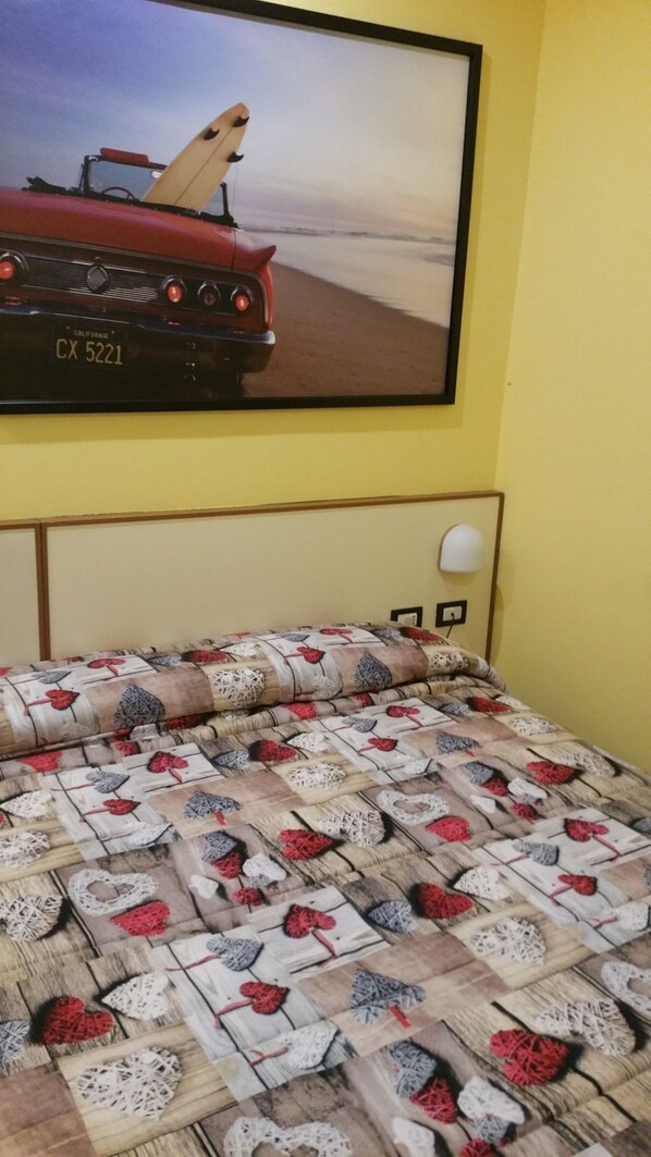 A bed with a patterned bedspread, a wall-mounted picture of a red car, and a bedside lamp. - Hotel Il Giardino (Pisa)