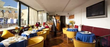 Un comedor con mesas dispuestas para comidas, un camarero atendiendo a los huéspedes, y un ventanal con vistas al exterior.