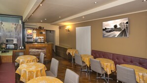 Un restaurante con mesas redondas cubiertas con manteles amarillos, una zona de bar con máquinas de café y un cuadro grande en la pared.