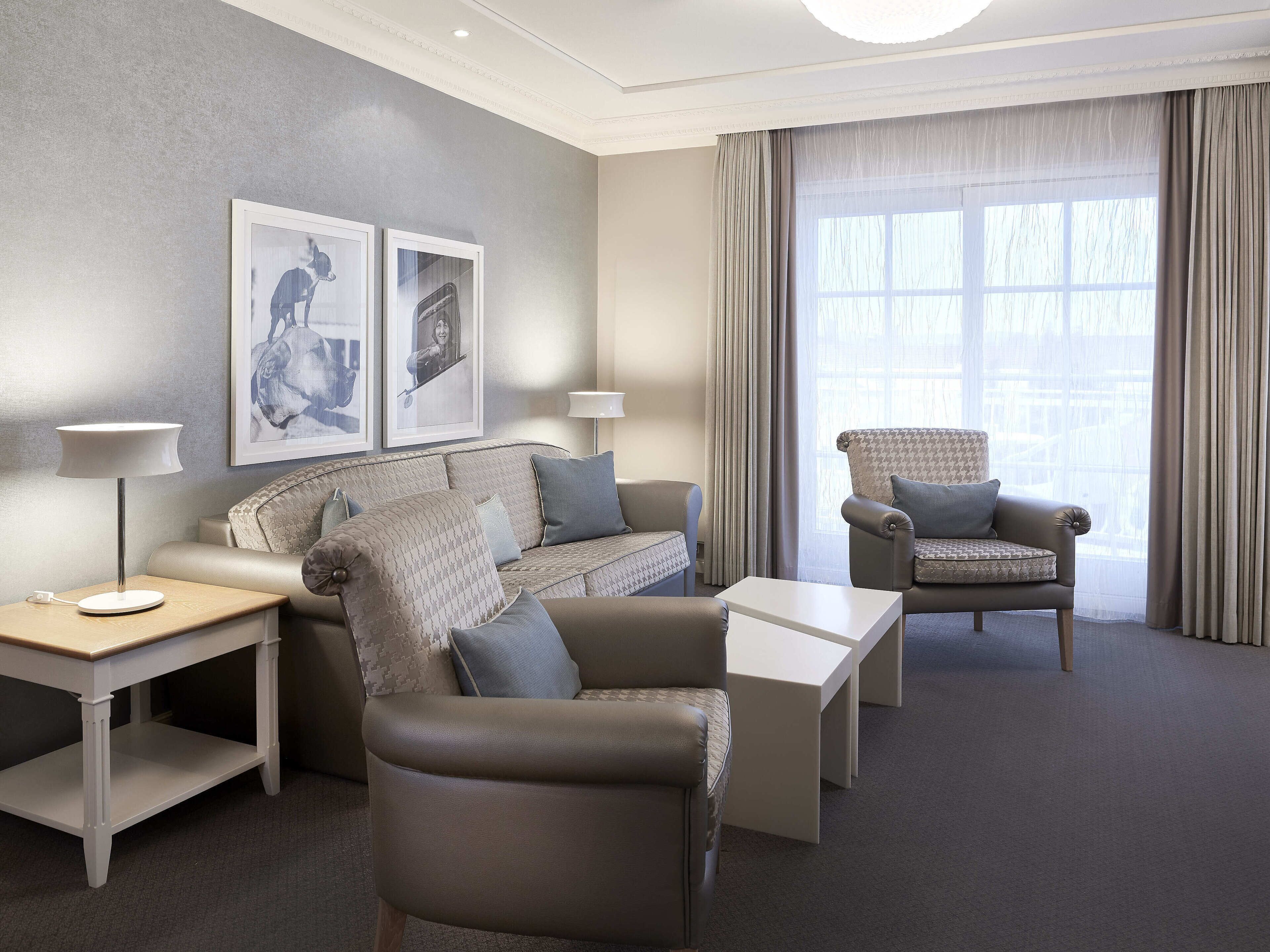 A modern hotel room with a grey patterned sofa, a leather armchair, a white side table, and a large window with sheer curtains.