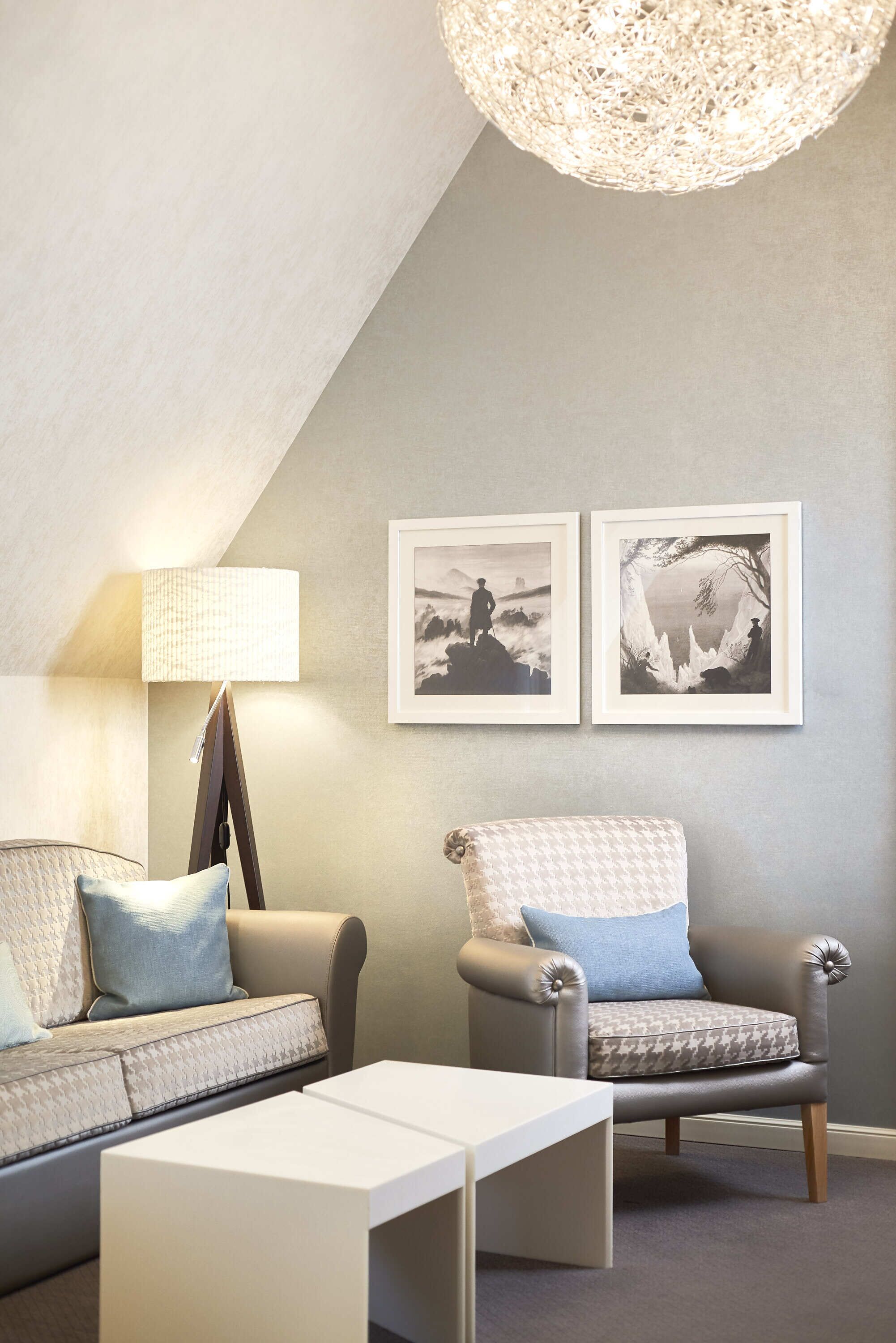 A modern living room with a sofa, armchair, and a coffee table. There are two framed pictures on the wall and a floor lamp.