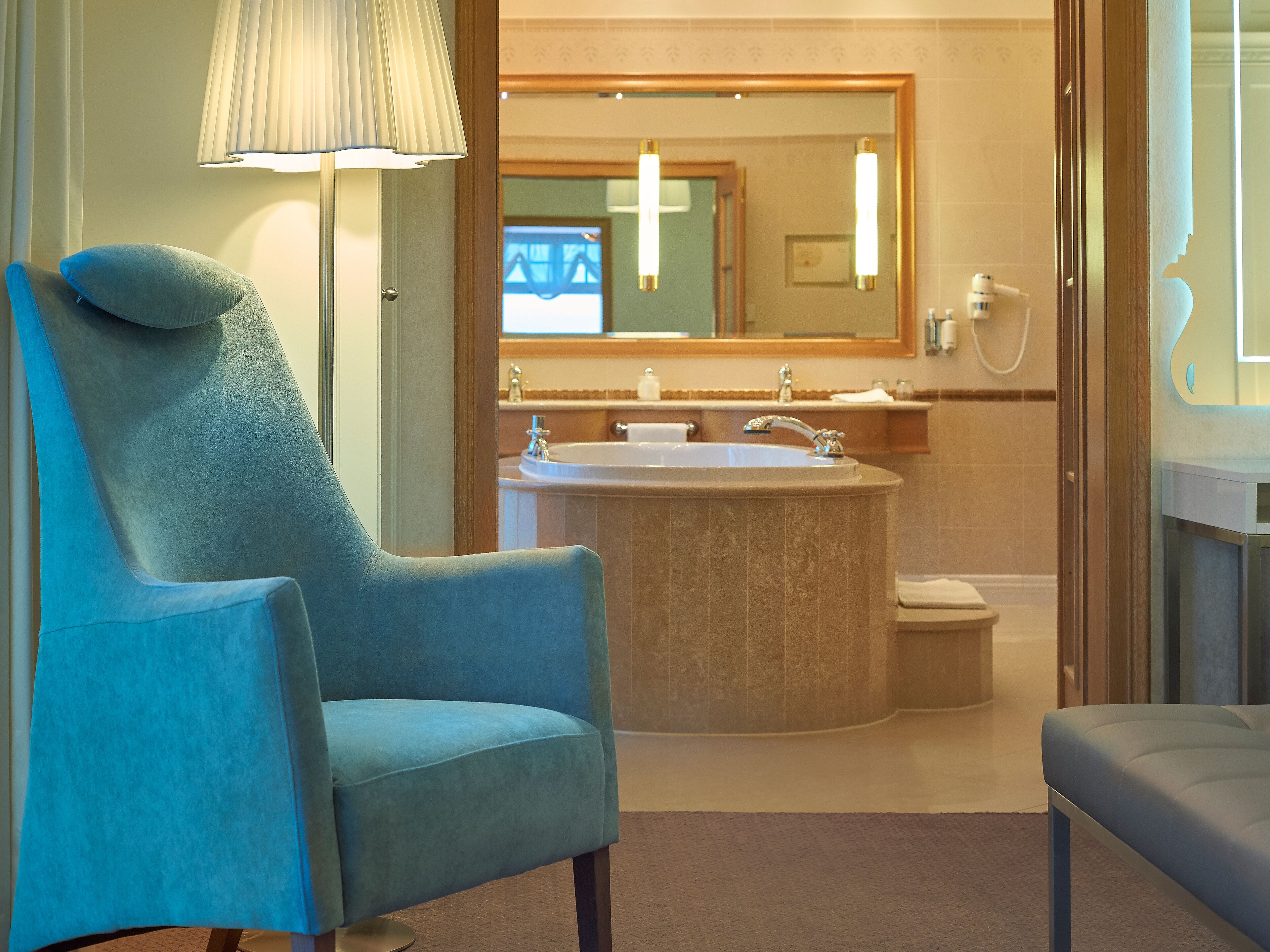 A modern bathroom with a large bathtub, a sink, and a mirror. There is a blue chair and a floor lamp.