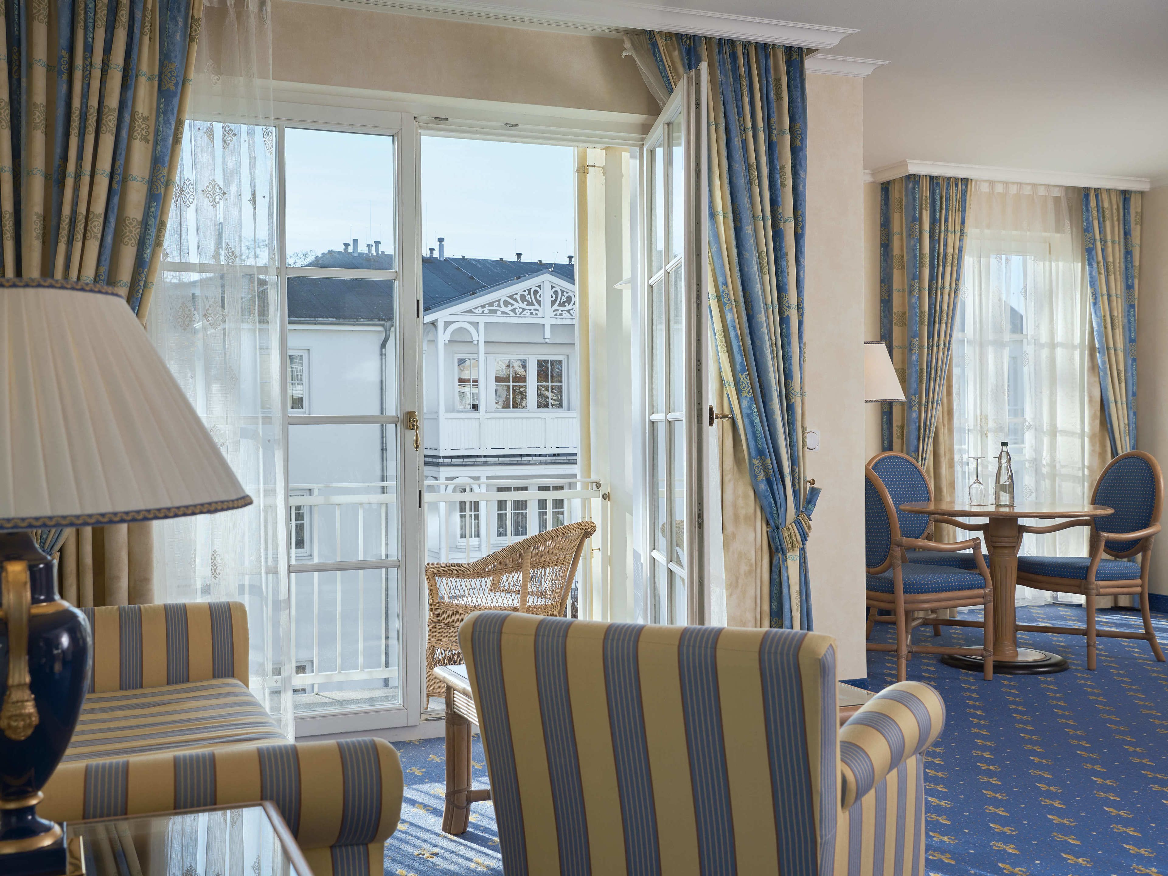 A hotel room with a balcony, striped armchairs, a dining table, and a view of a building.